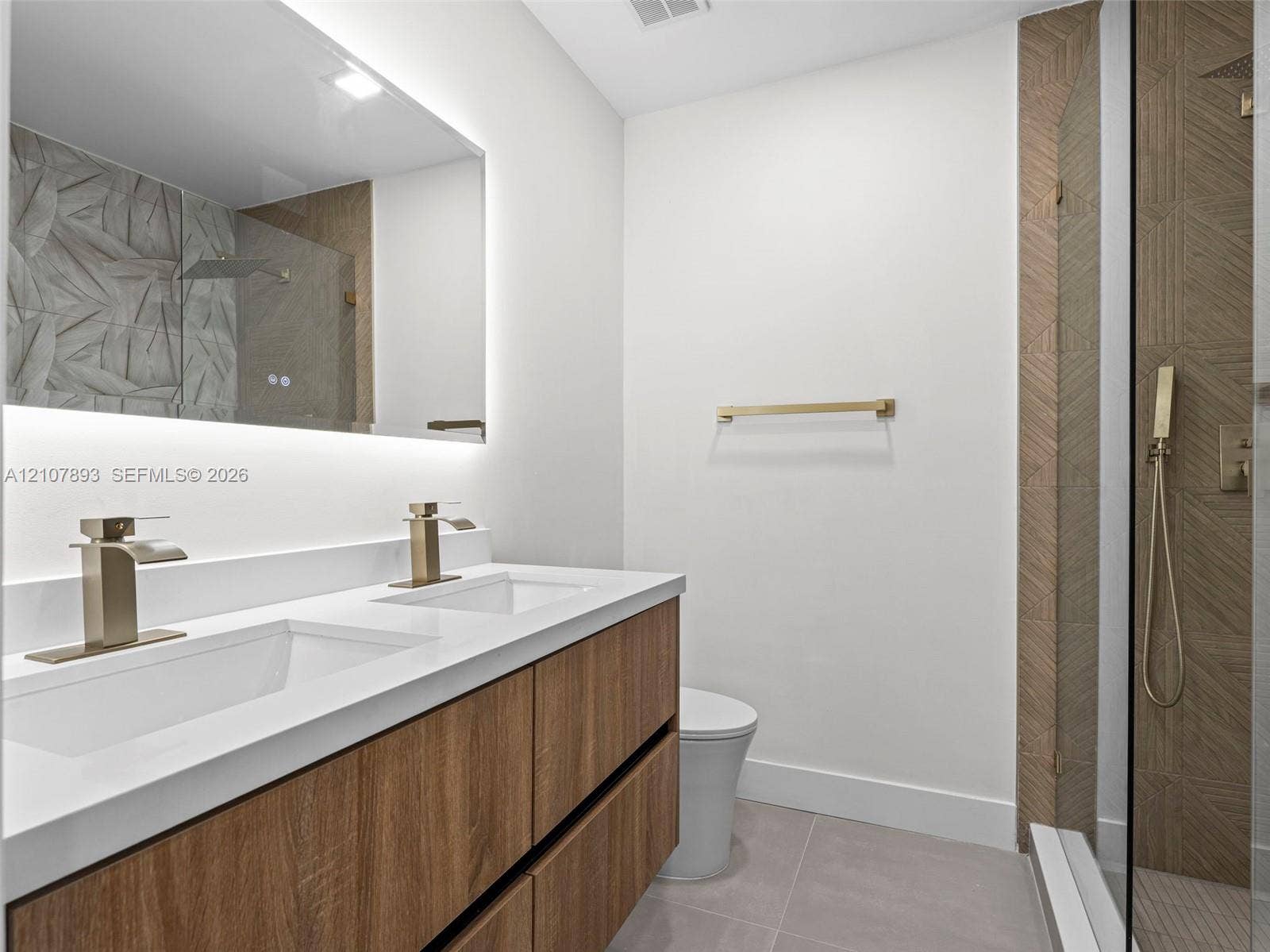 610 NE 164th Terrace, Miami, FL 33162 Floating wood-finish double vanity with white countertop, integrated rectangular sinks, brass-toned fixtures, LED-backlit mirror, and glass-enclosed shower with patterned tile surround