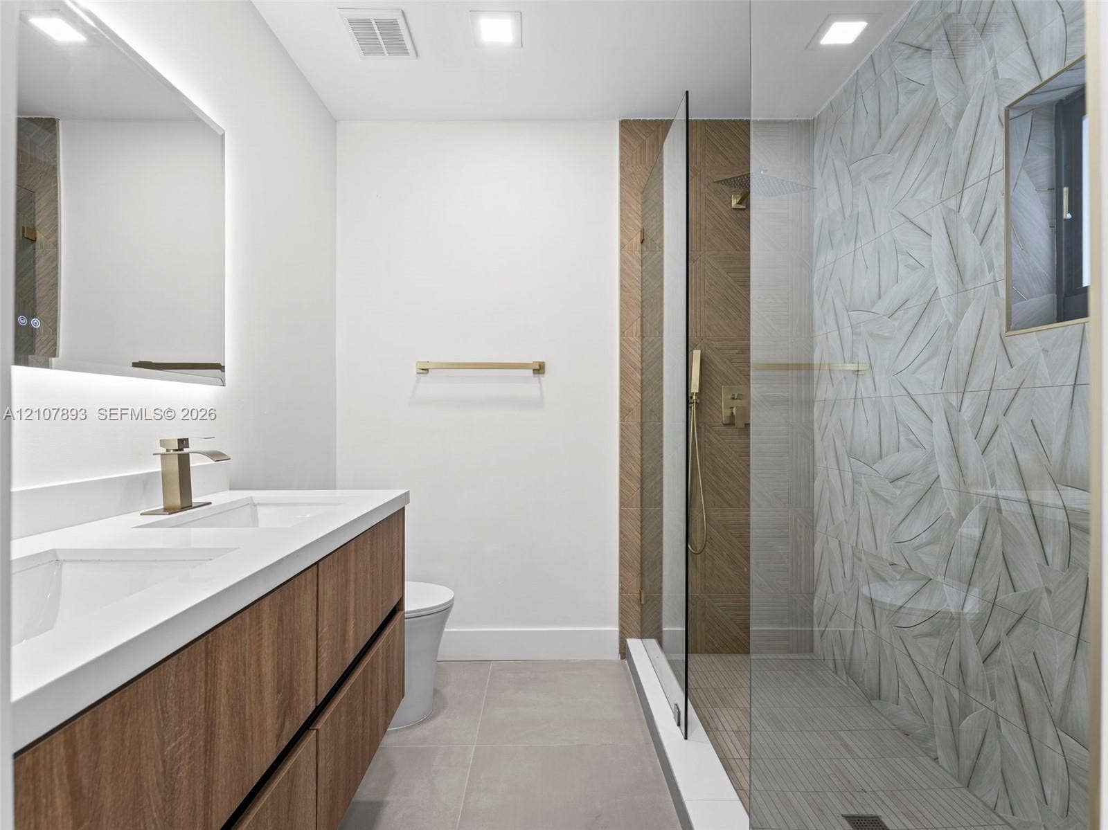 610 NE 164th Terrace, Miami, FL 33162 Double vanity with quartz countertop, wood-finish cabinetry, frameless glass-enclosed shower featuring textured leaf-patterned wall tile, and matte gold-tone fixtures