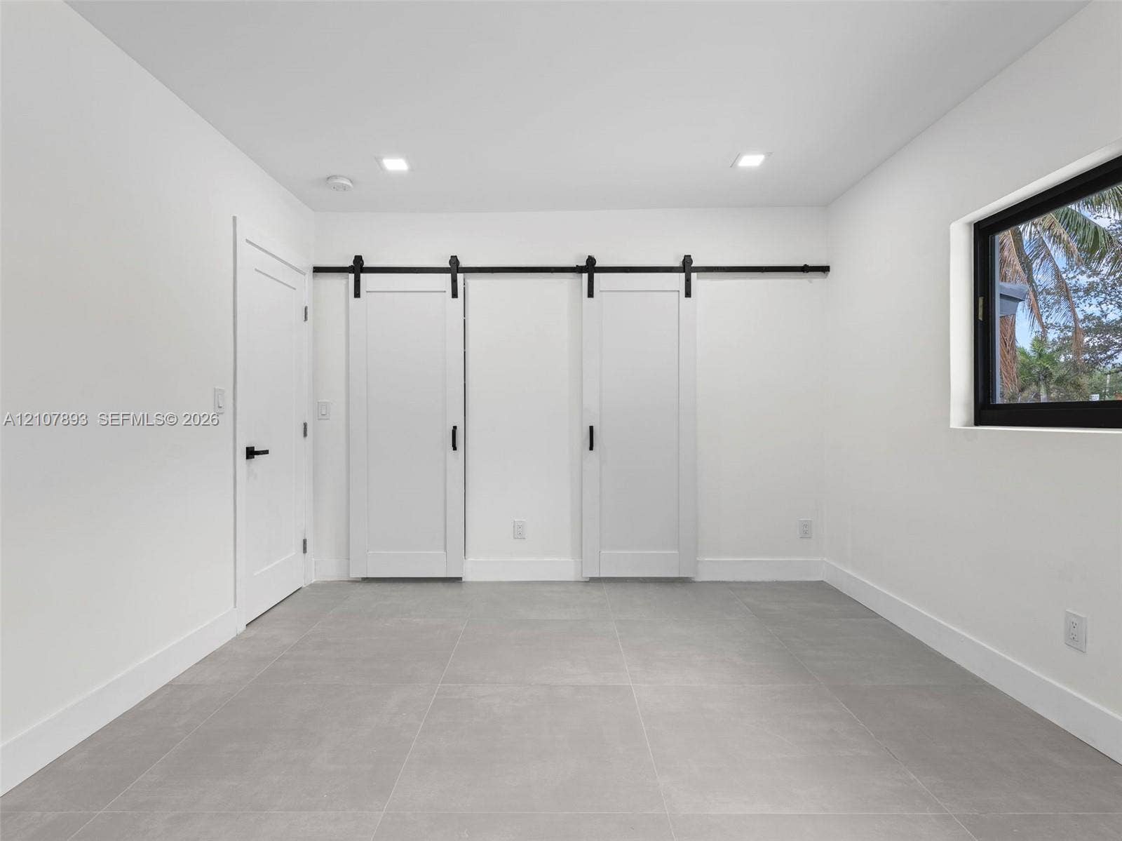 610 NE 164th Terrace, Miami, FL 33162 Interior room featuring large-format ceramic tile flooring, dual white sliding barn doors with matte black hardware, recessed ceiling lighting, and a window with a black frame