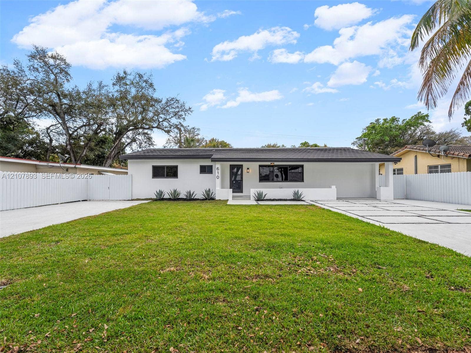 610 NE 164th Terrace, Miami, FL 33162 Single-story exterior featuring white stucco siding, a low-slope roof, expansive front lawn, concrete paver driveway with integrated turf, and full-perimeter privacy fencing