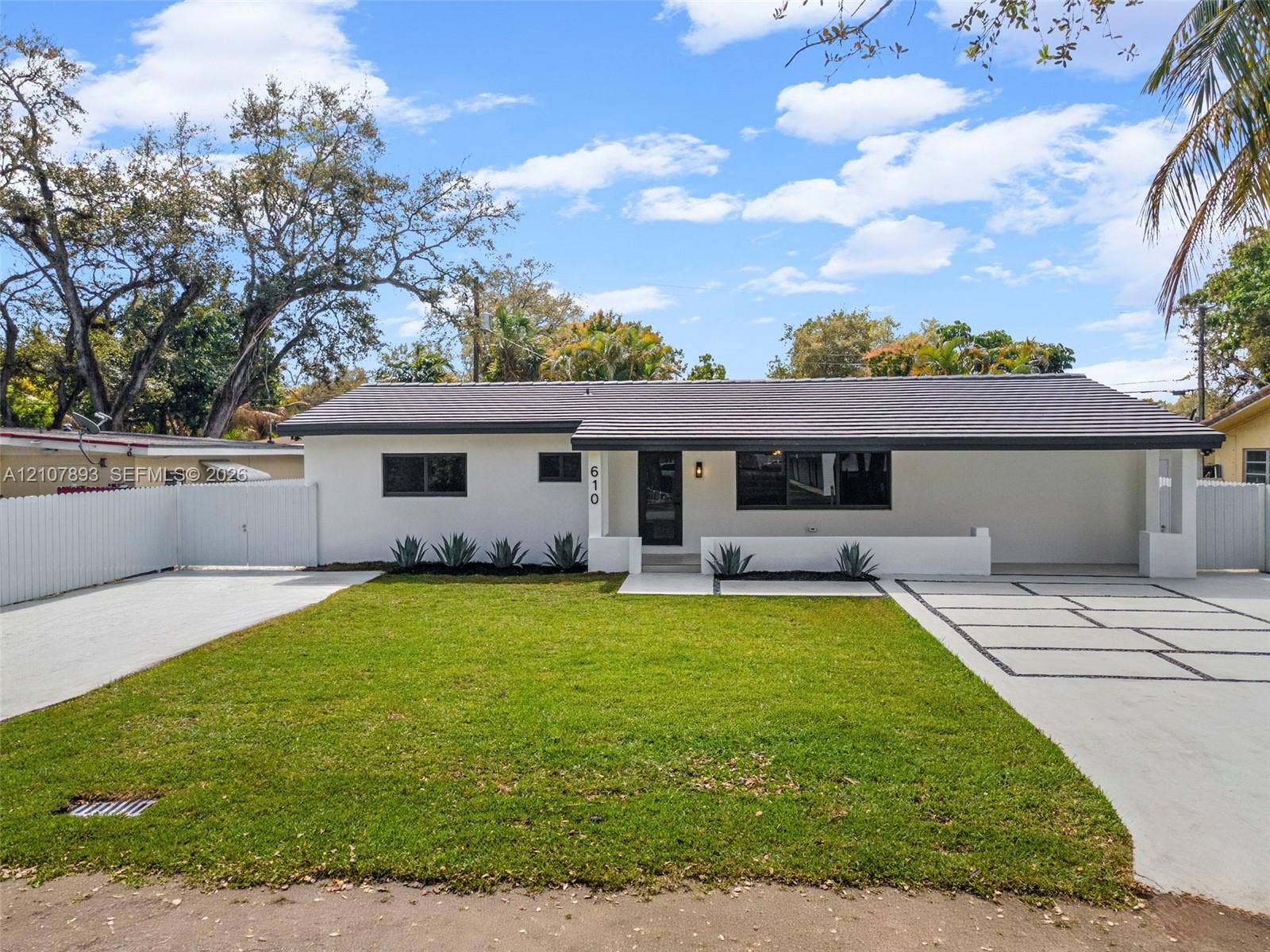 610 NE 164th Terrace, Miami, FL 33162 Single-story exterior featuring white stucco finish, dark-toned roof, paved geometric driveway, white perimeter fencing, and manicured lawn
