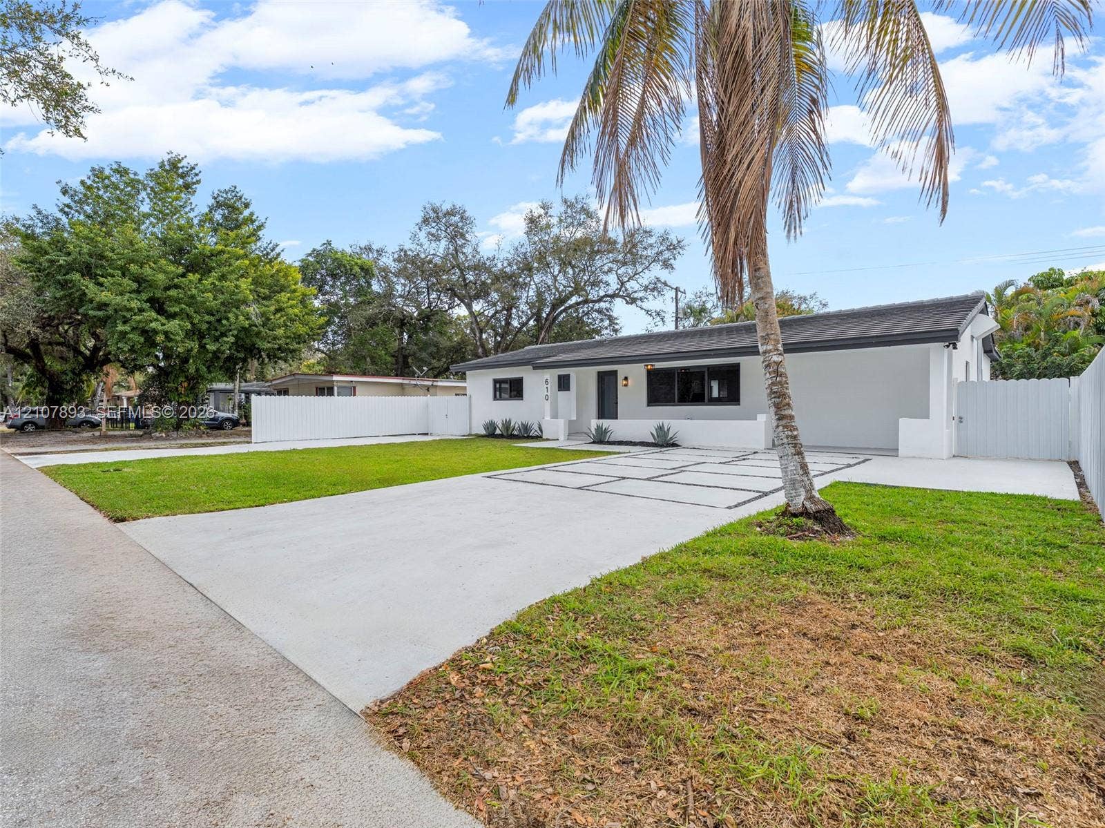 610 NE 164th Terrace, Miami, FL 33162 Single-story exterior featuring a painted stucco finish, attached carport, grid-patterned concrete driveway, white perimeter fencing, and established palm trees