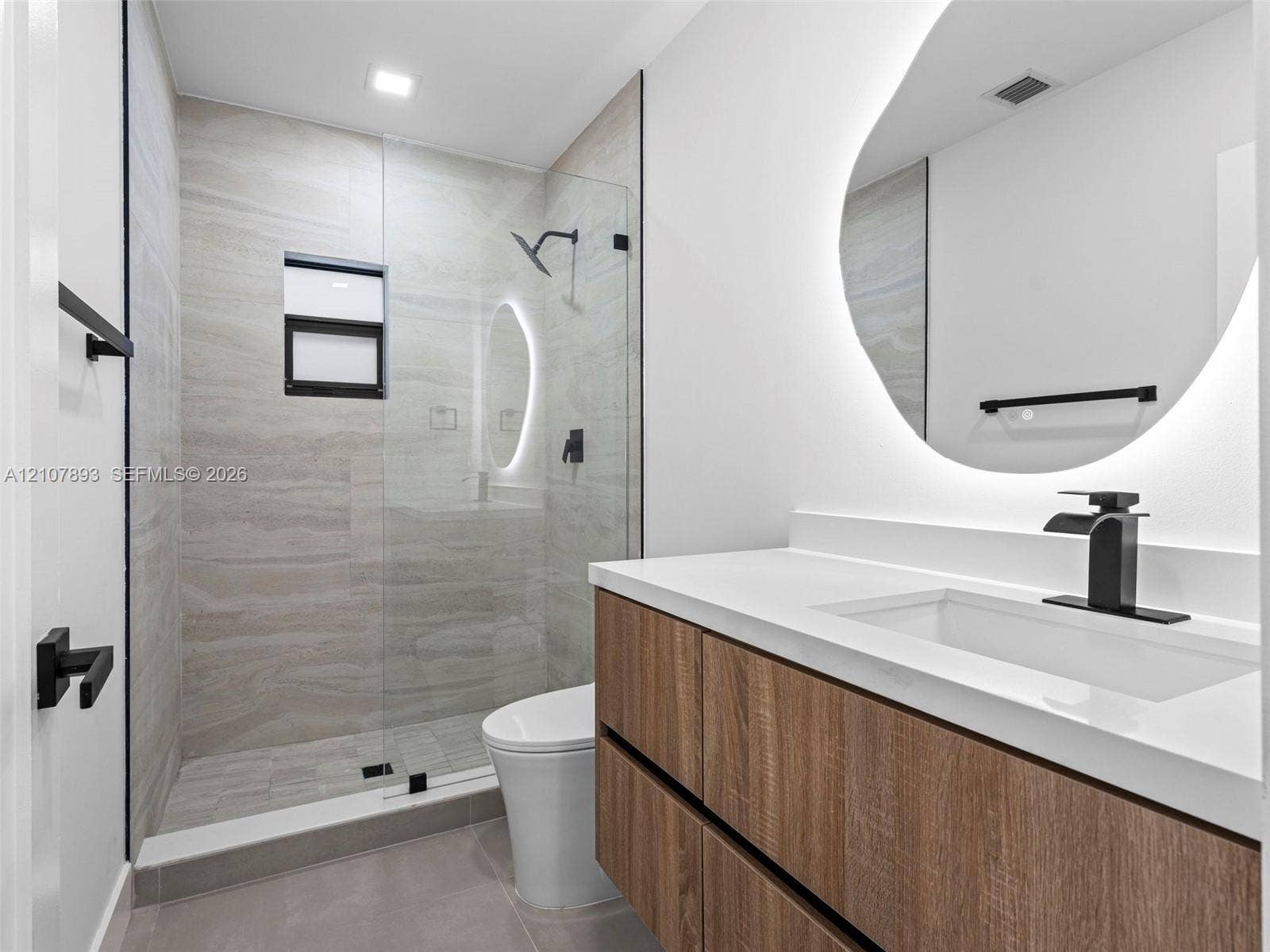 610 NE 164th Terrace, Miami, FL 33162 Wood-finish floating vanity with a white countertop, integrated rectangular sink, and matte black faucet, featuring a wall-mounted back-lit mirror, glass-enclosed shower with horizontal-patterned tile surround, and matte black hardware
