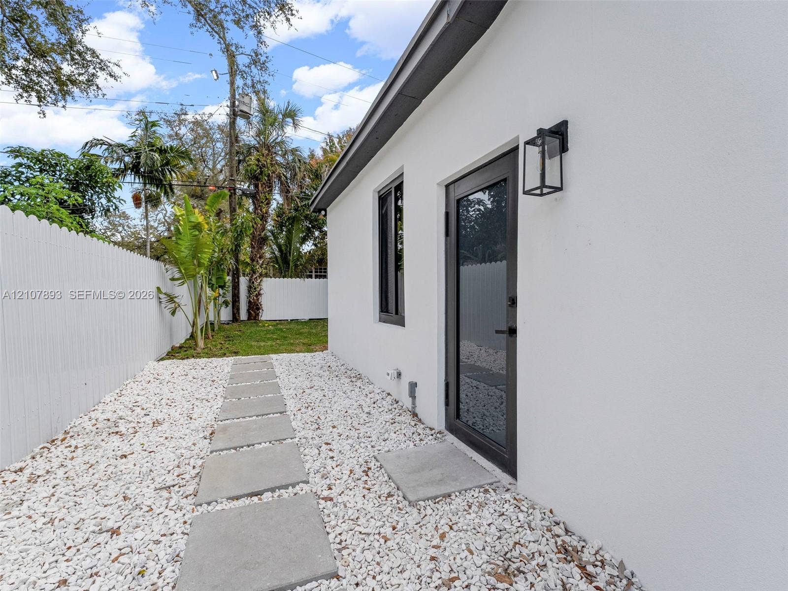 610 NE 164th Terrace, Miami, FL 33162 Stucco exterior featuring a dark-framed glass door, exterior wall-mounted lantern lighting, white privacy fencing, and a stone-paver walkway set in gravel