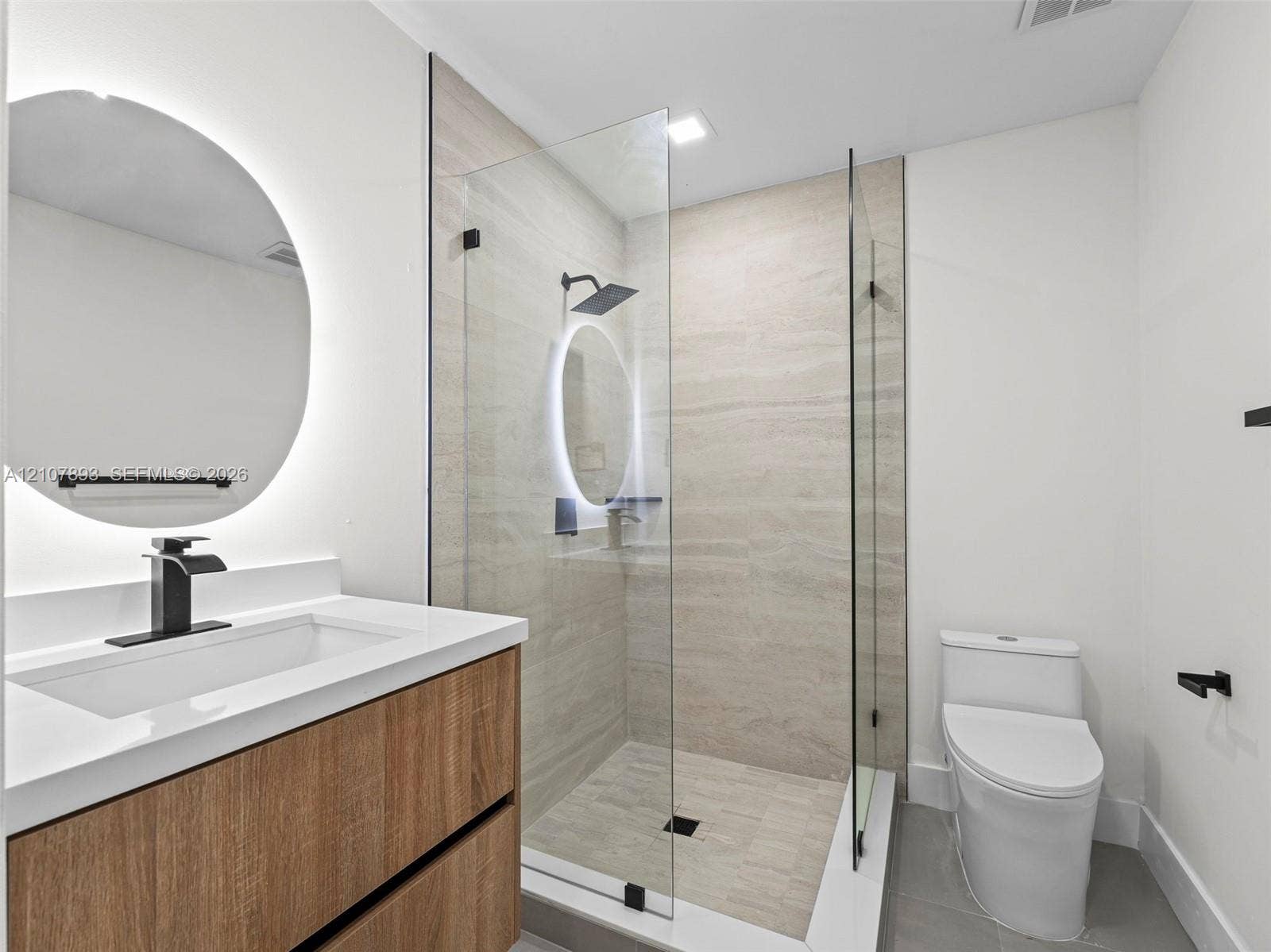 610 NE 164th Terrace, Miami, FL 33162 Floating vanity with wood-finish cabinetry, quartz countertop, illuminated circular mirror, glass-enclosed walk-in shower with matte black fixtures, and dual-flush toilet