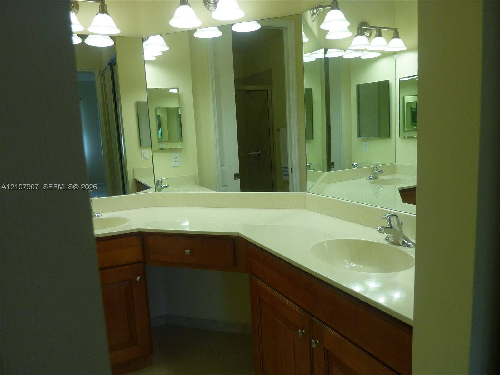 L-shaped vanity featuring wood-finish cabinetry, dual integrated sinks, light-toned solid surface countertops, and multi-bulb vanity lighting
