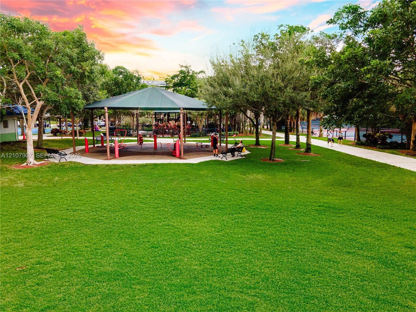 Community park space featuring a large manicured lawn, shaded pavilion, mature landscaping, paved pathways, and outdoor seating