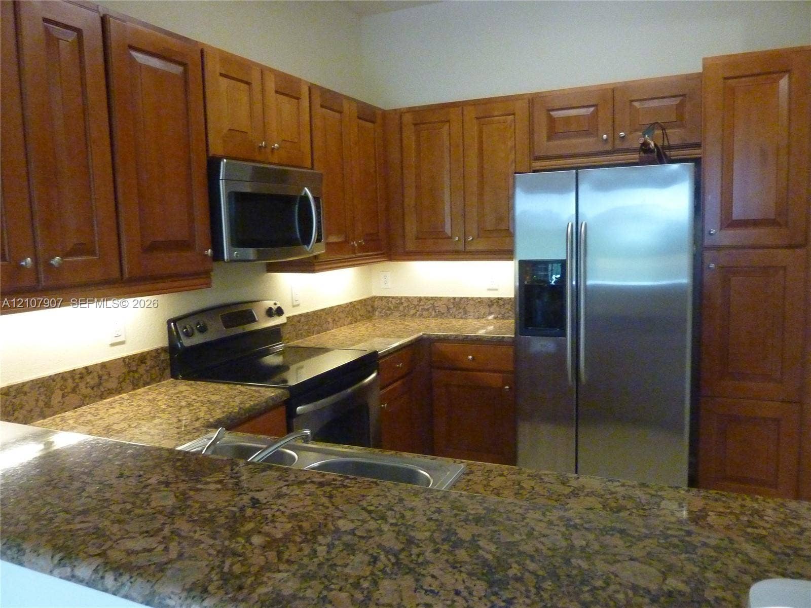 Kitchen featuring wooden cabinetry, stone-surface countertops, stainless steel appliances, a double-basin sink, and under-cabinet lighting