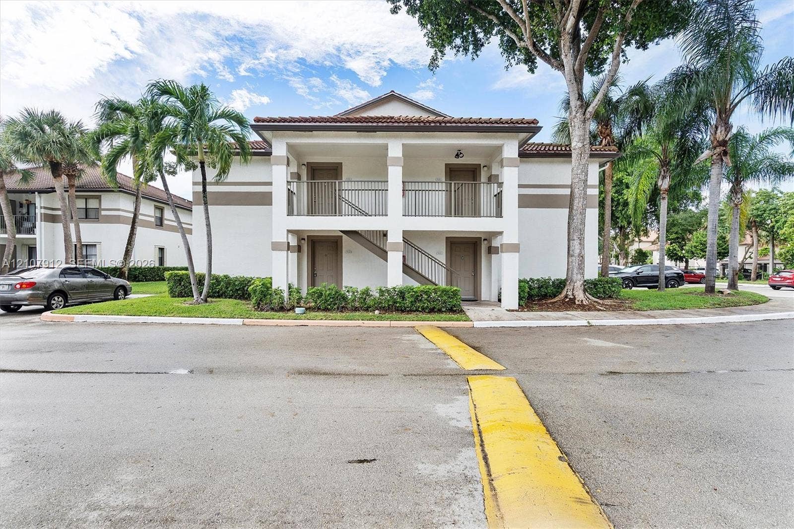 Two-story exterior featuring a stucco facade, covered balconies, metal railings, and a tiled roof, accented by mature palm trees and manicured shrubbery