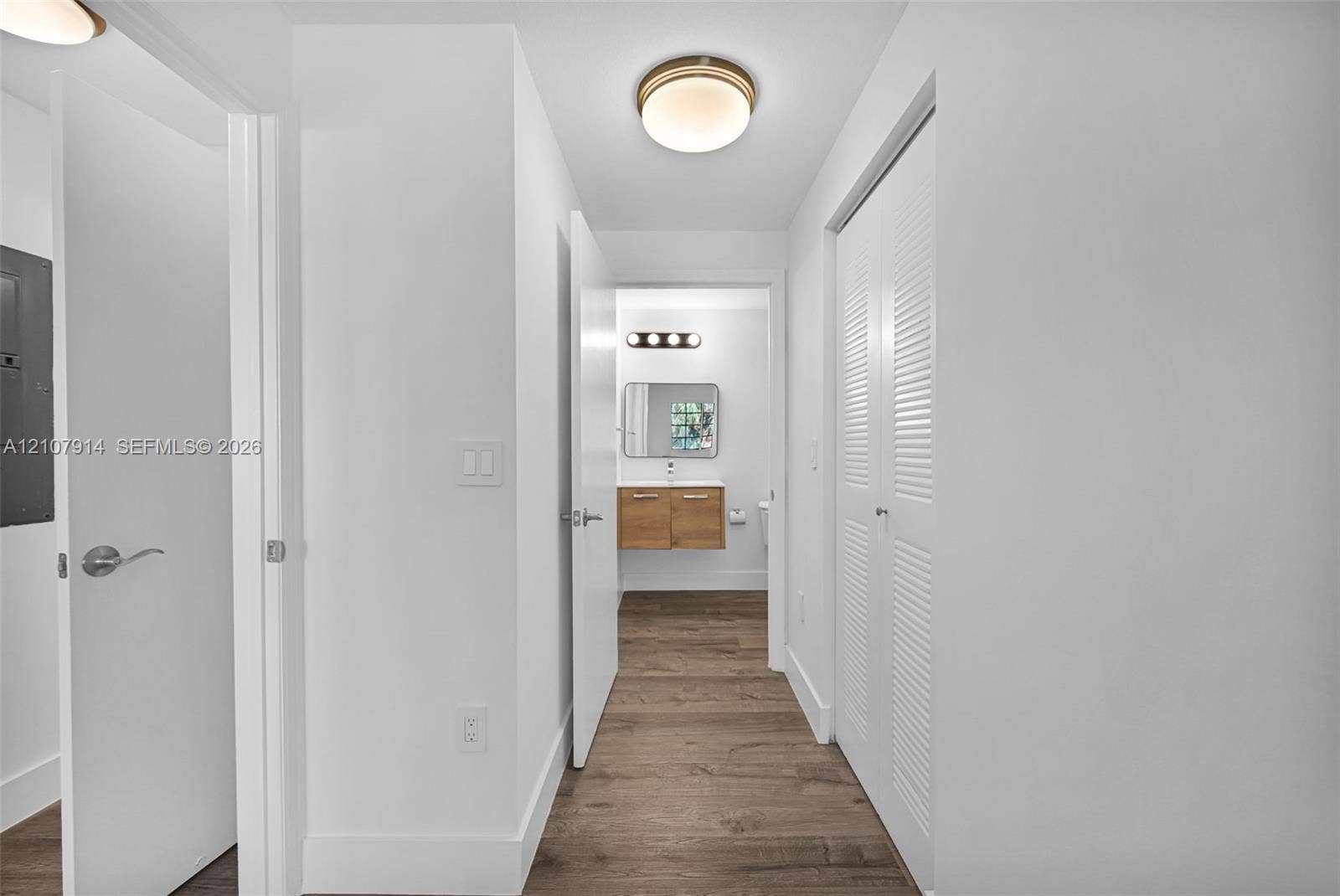 7340 NW 114th Avenue, #205, Doral, FL 33178 Hallway featuring wood-finish flooring, white louvered closet doors, and a flush-mount ceiling light fixture, opening into a space with a wall-mounted vanity, rectangular mirror, and four-light vanity bar