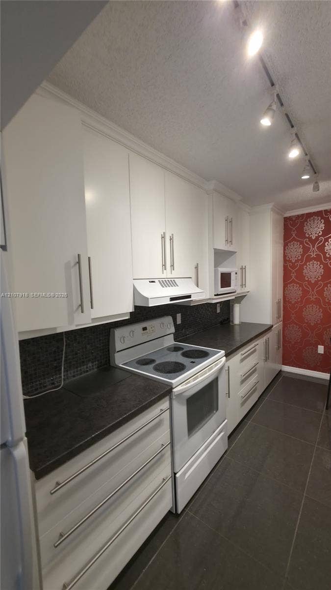 Kitchen featuring flat-panel white cabinetry, dark countertops, a dark mosaic tile backsplash, built-in track lighting, and ceramic tile flooring