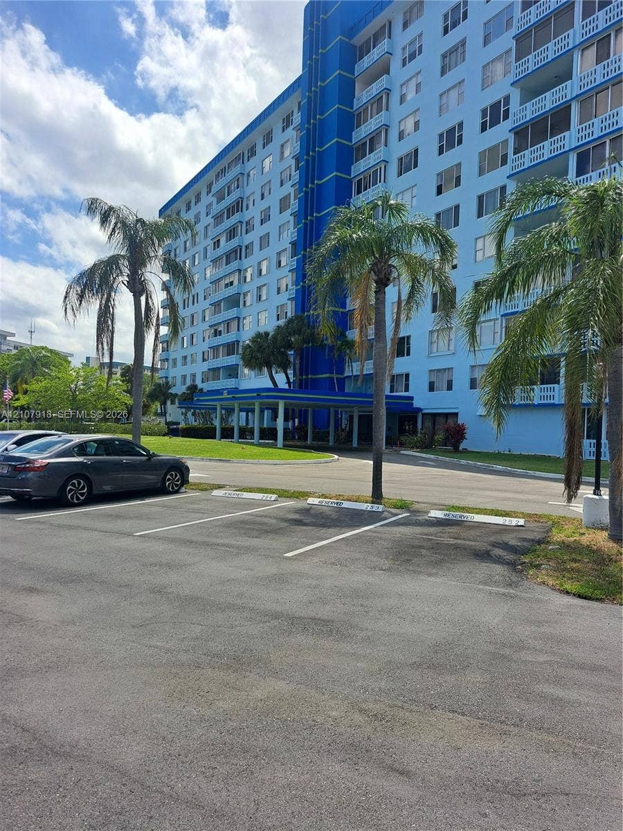 Multi-story residential building featuring a covered arrival portico, private balconies with integrated railings, asphalt parking surface, and mature palm landscaping