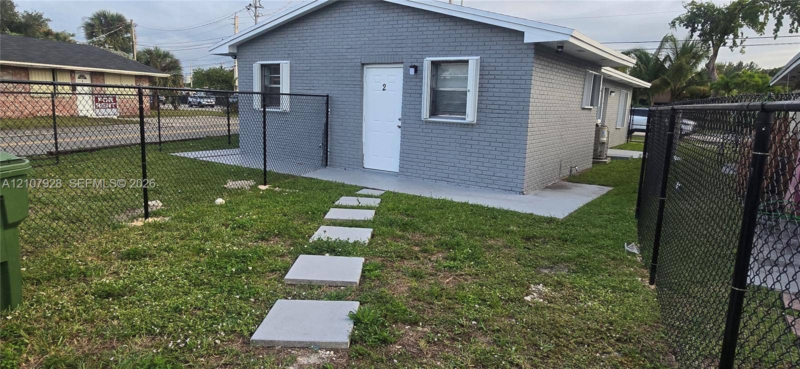 815 Foster Road, #2, Hallandale Beach, Hallandale Beach, FL 33009 Painted brick exterior featuring a concrete entry pad, square paver walkway, and black chain-link perimeter fencing