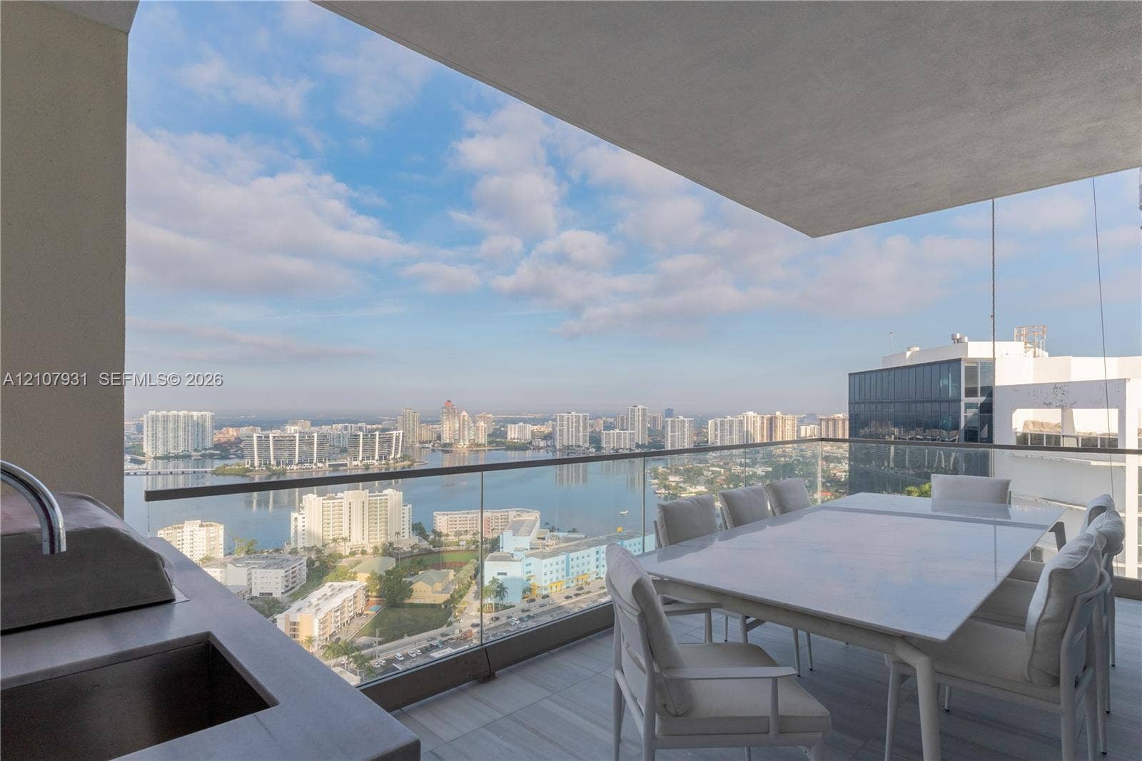 Covered outdoor balcony featuring a built-in outdoor kitchen with sink, glass panel railing, and tile flooring