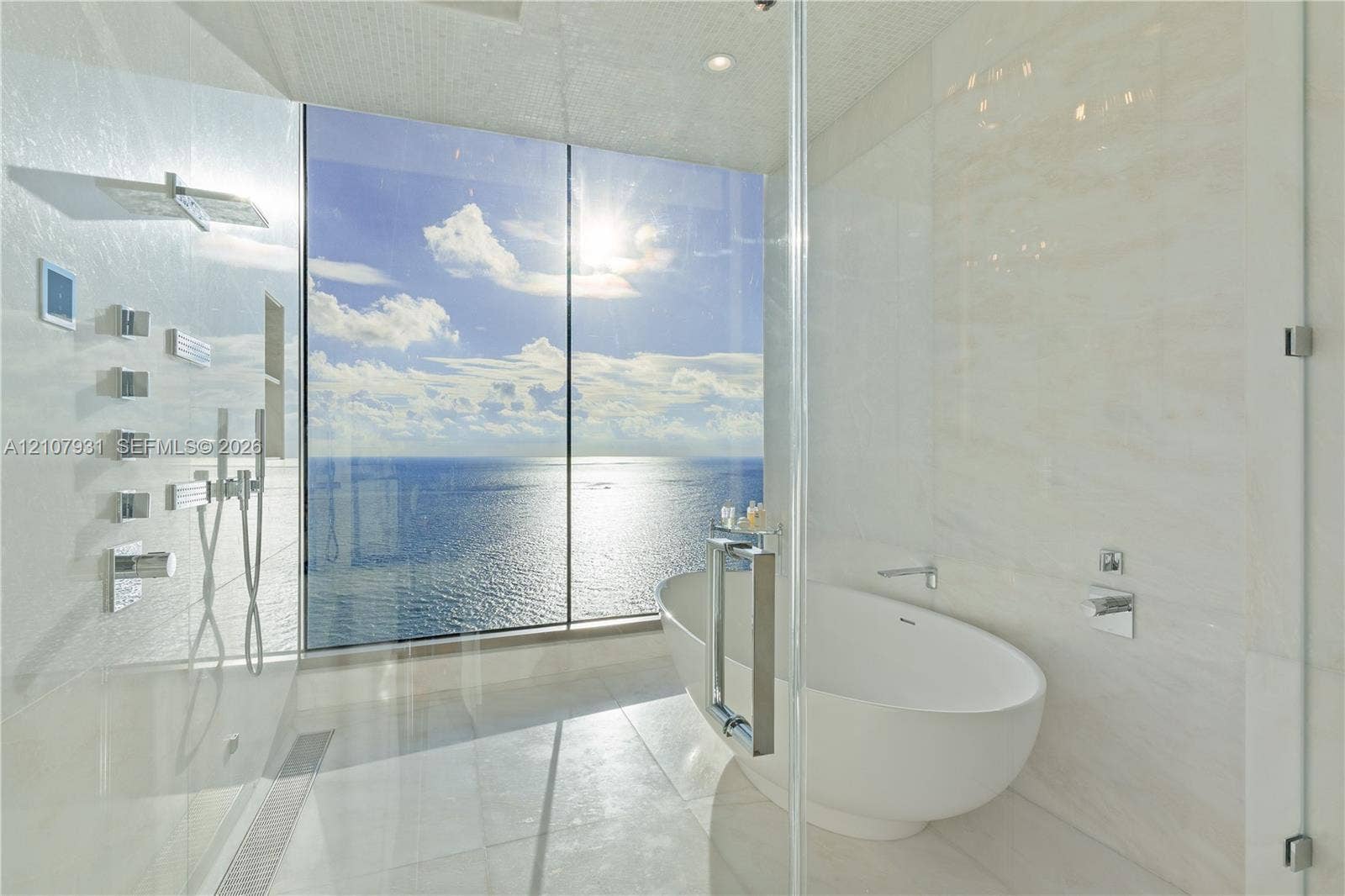 Floor-to-ceiling glass paneling, freestanding soaking tub, walk-in shower with multi-head wall-mounted shower system, and light-toned tile flooring