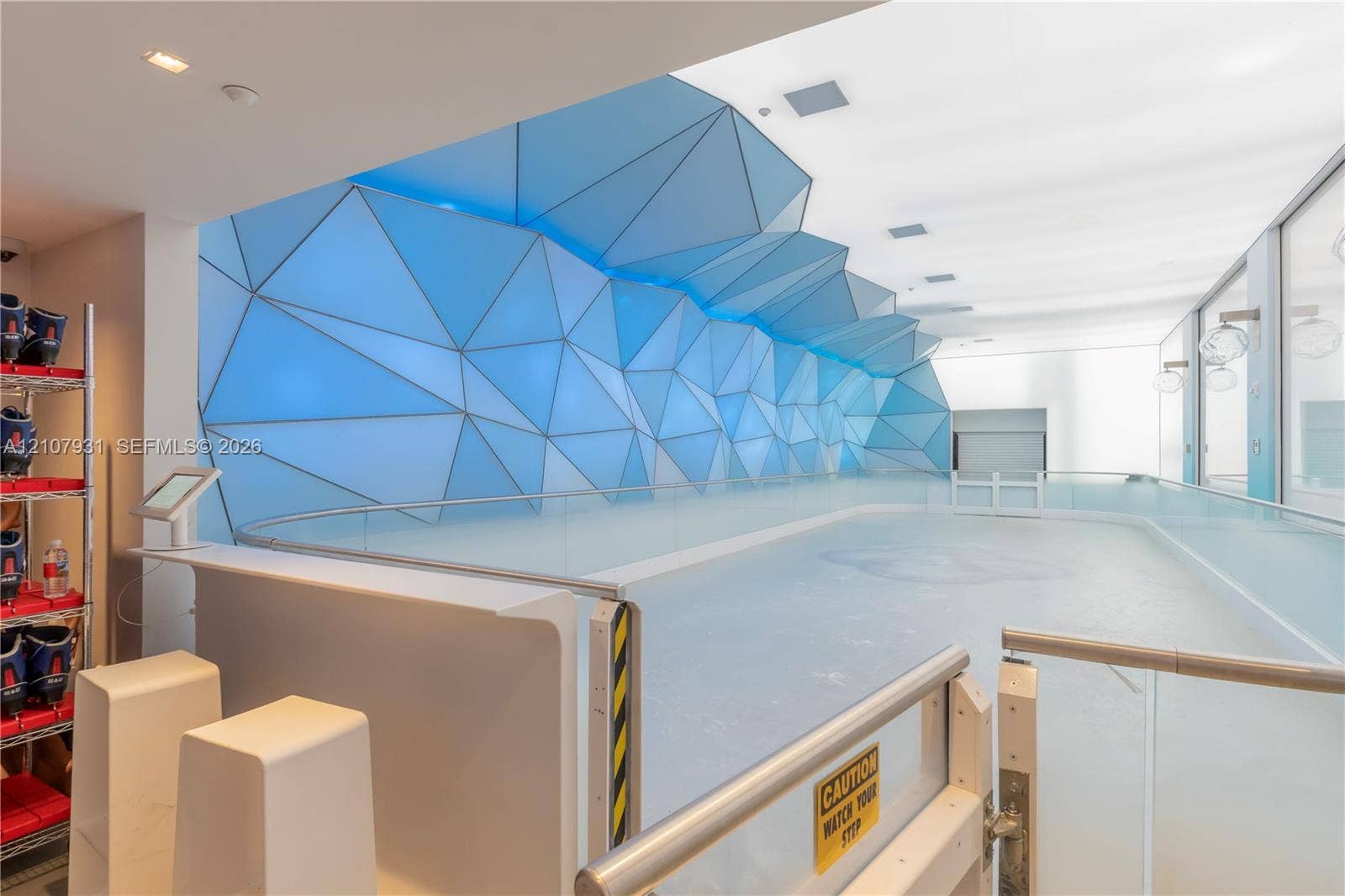 Indoor ice skating rink featuring a geometric-patterned illuminated feature wall, frameless glass panel railings, and white ceiling with recessed lighting