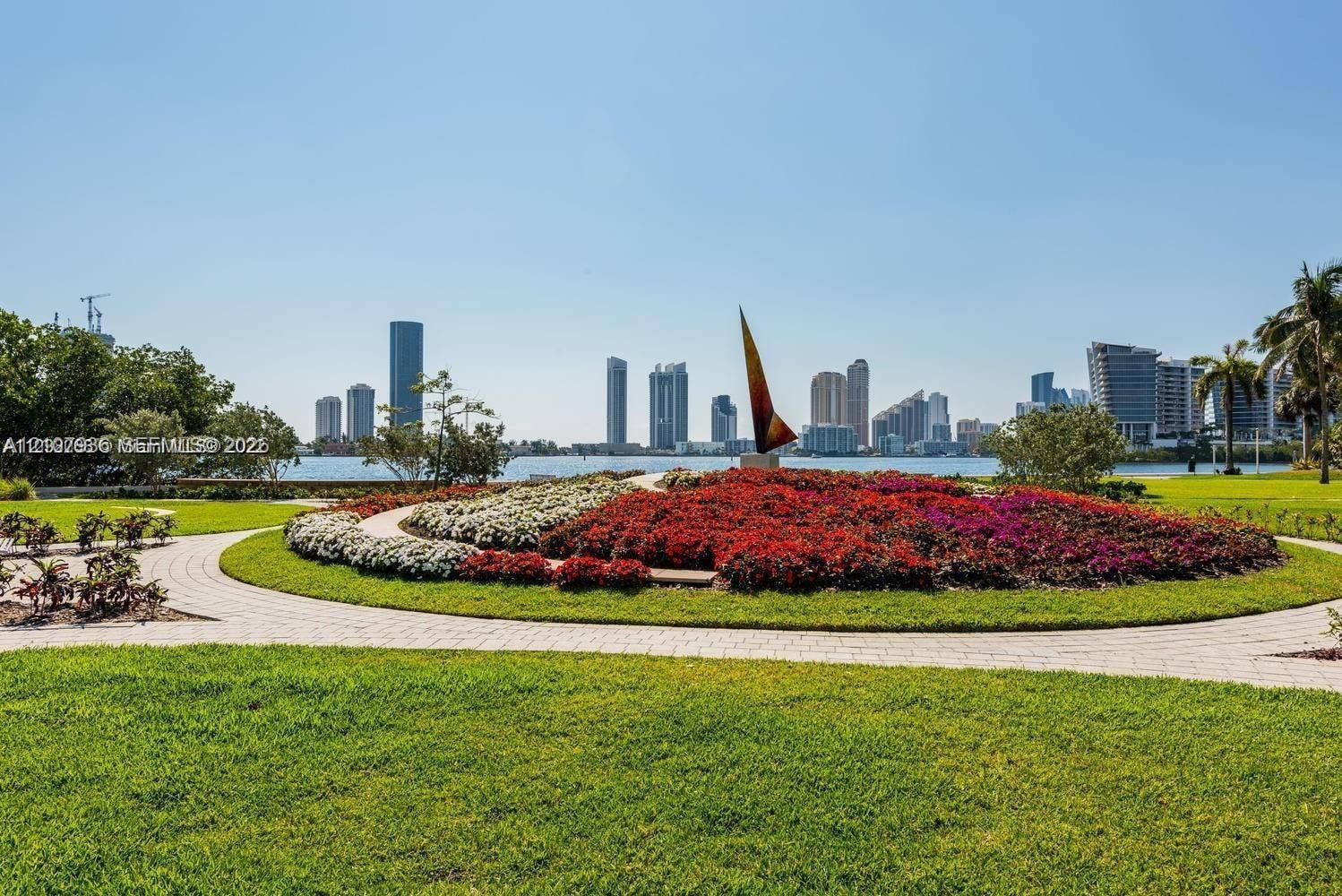 3370 Hidden Bay Drive, #510, Aventura, FL 33180 Manicured lawn featuring a circular stone paver path, expansive floral landscaping, decorative sculpture, and water frontage with a cityscape backdrop