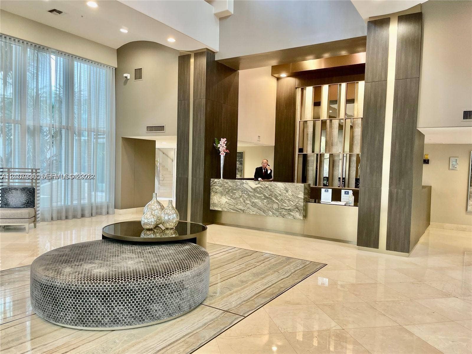3370 Hidden Bay Drive, #510, Aventura, FL 33180 Double-height lobby featuring floor-to-ceiling windows, polished stone flooring, and a decorative stone-front reception desk accented with metallic paneling and dark wood-finish columns