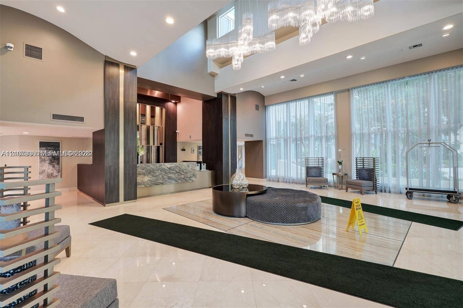 3370 Hidden Bay Drive, #510, Aventura, FL 33180 Double-height ceiling lobby featuring stone-finish flooring, a tiered crystal chandelier, floor-to-ceiling windows, and a reception desk with stone-finish facade