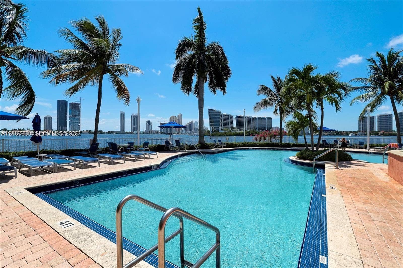3370 Hidden Bay Drive, #510, Aventura, FL 33180 Swimming pool featuring tiled border, metal handrails, and paved deck surrounded by palm trees with waterfront skyline backdrop