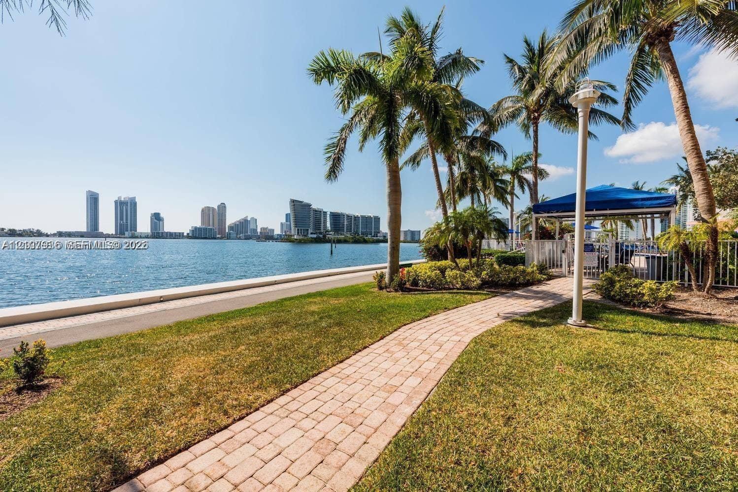 3370 Hidden Bay Drive, #510, Aventura, FL 33180 Waterfront grounds featuring a brick paver pathway, mature palm trees, coastal landscaping, and perimeter fencing