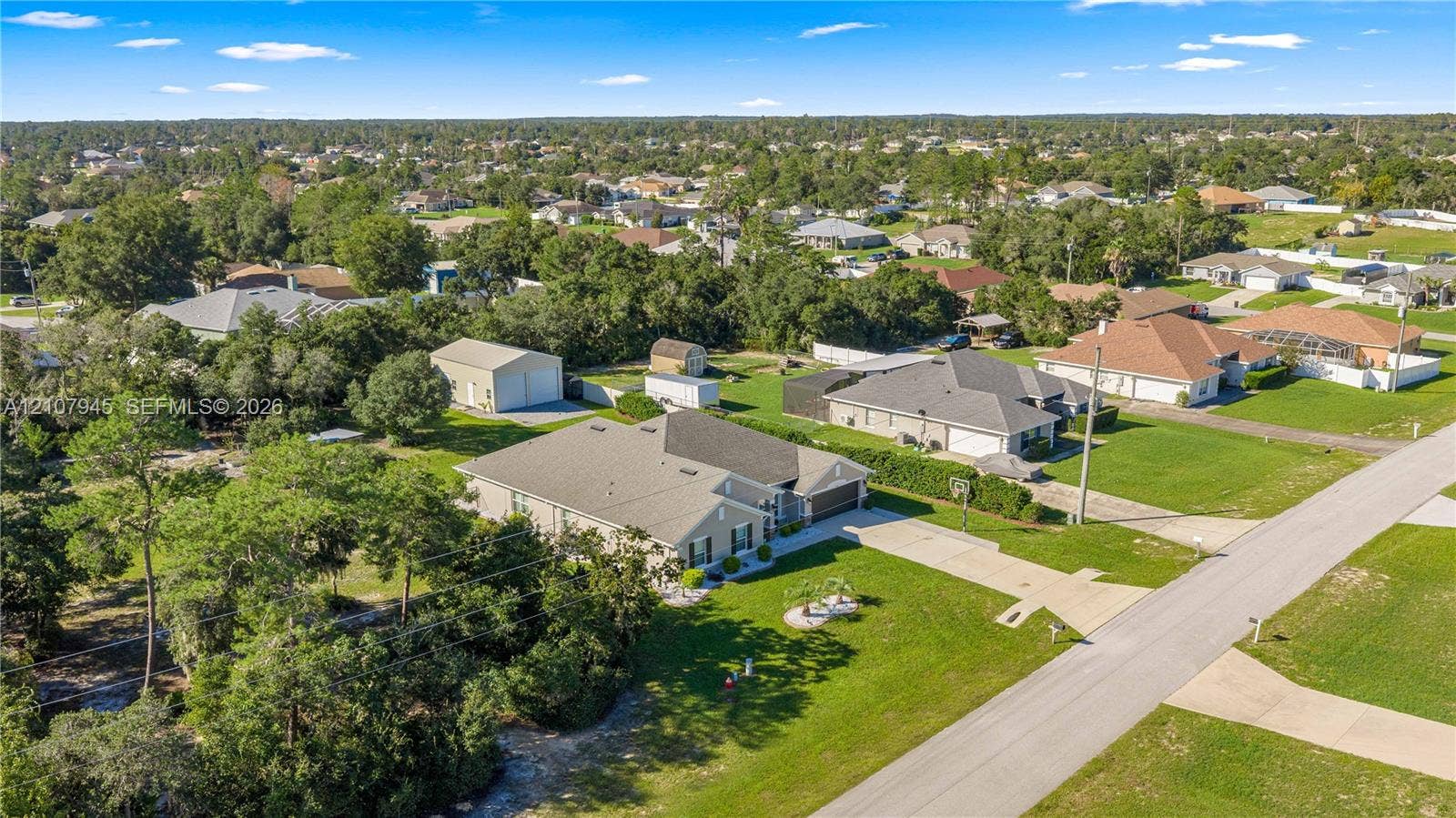 Aerial view of a property featuring a single-story residence, a multi-car paved driveway, a detached metal-sided storage structure, and expansive grounds with mature trees