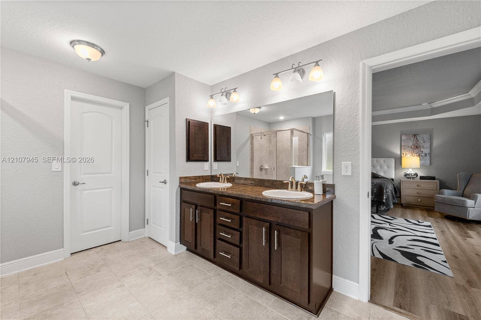 Dual-sink vanity with dark wood cabinetry, stone-finish countertop, full-width vanity mirror, and tile flooring