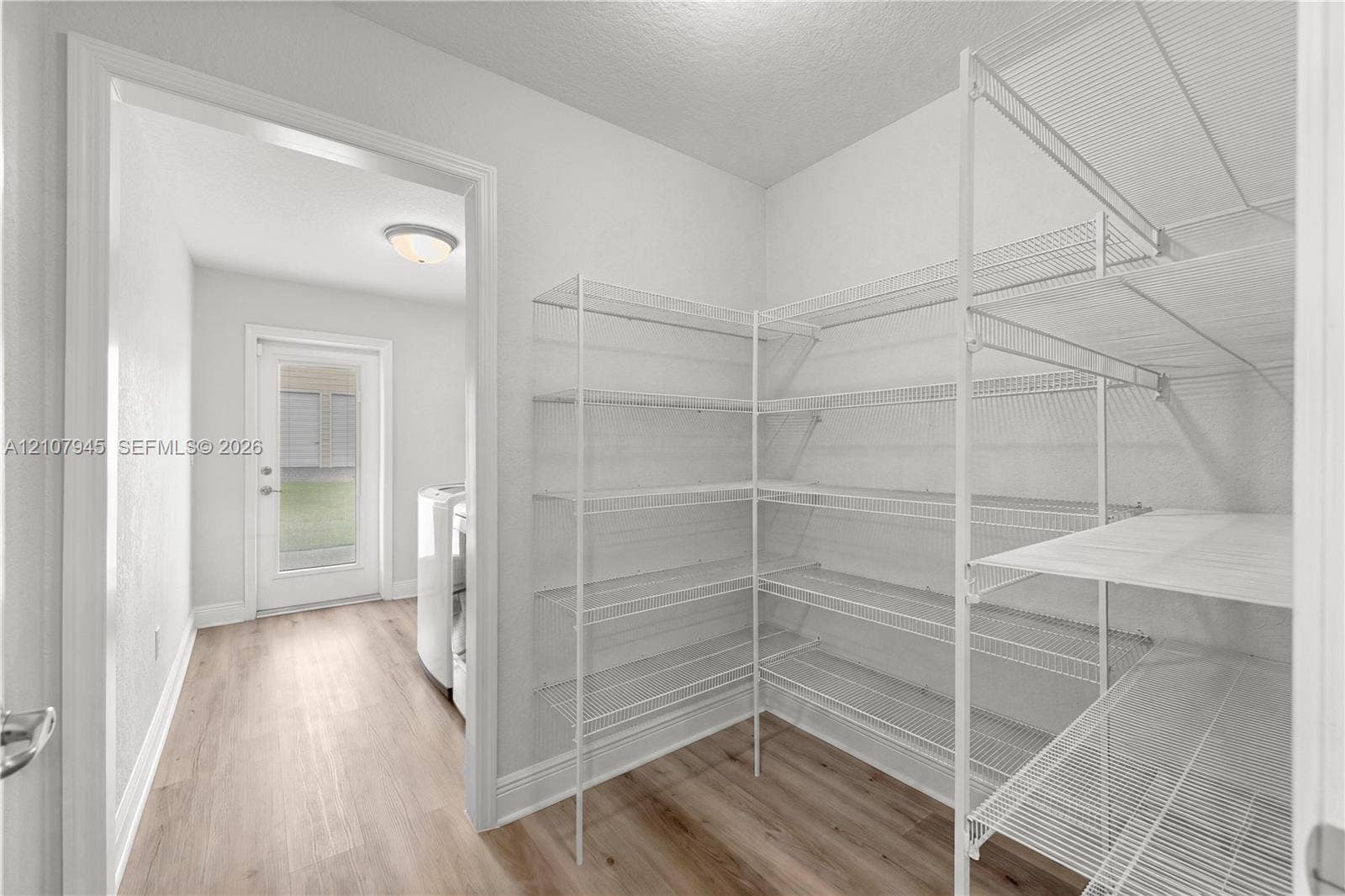 Storage room featuring white wire shelving and wood-finish flooring, with a doorway leading to a utility space with a glass-paneled exterior door