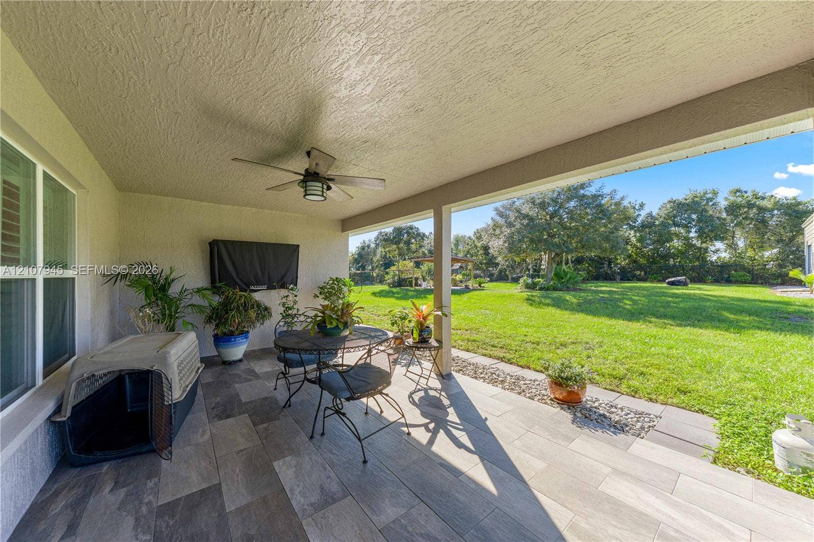 Covered patio featuring ceramic tile flooring, a ceiling-mounted fan, and a built-in television wall mount, overlooking a spacious lawn with mature tree line