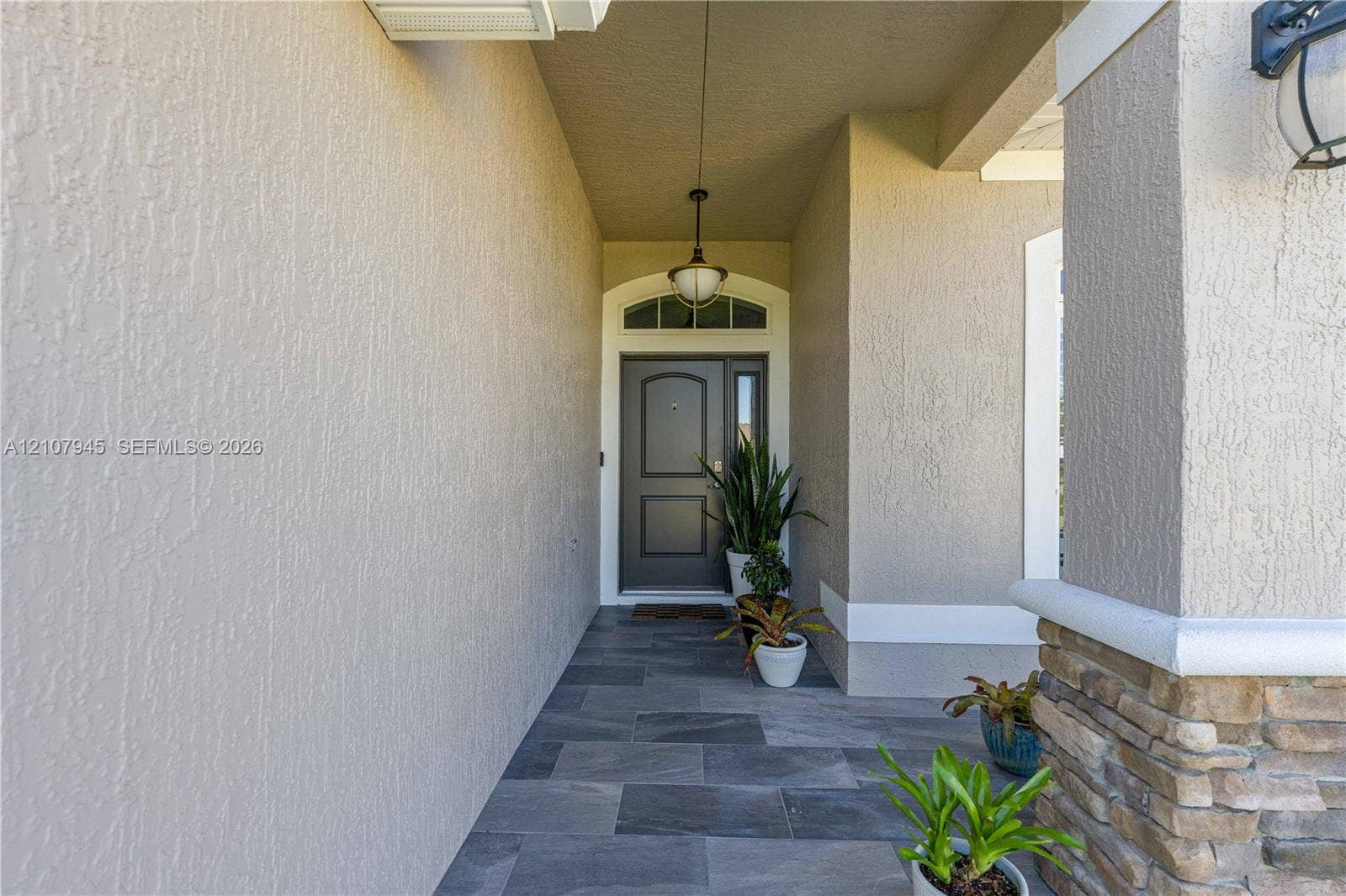Covered entryway featuring a stone veneer column base, tile flooring, and a single-panel front door with an arched transom window and integrated sidelight
