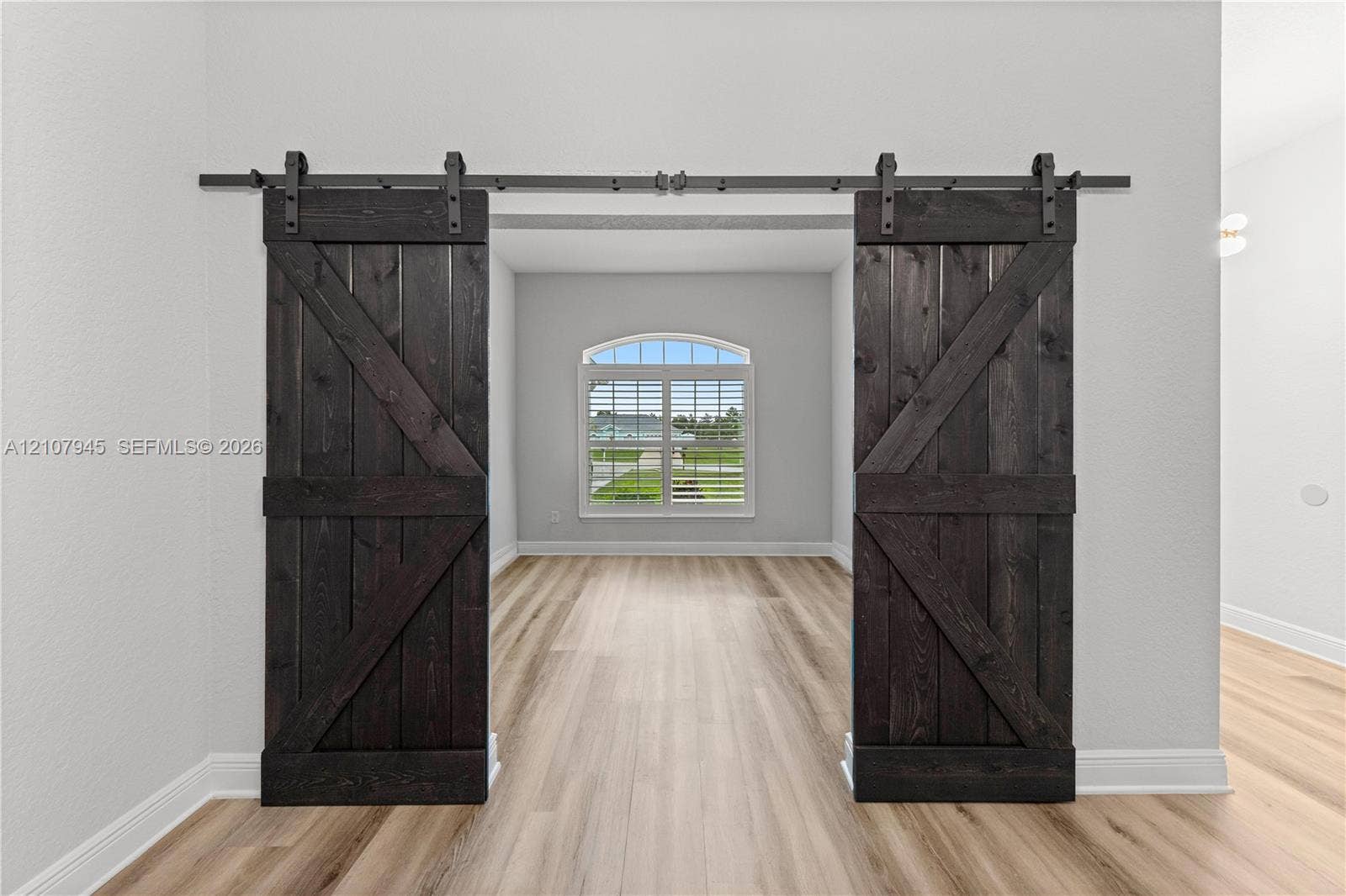 Dark-stained wood barn doors featuring matte black hardware, arched window with shutters, and wood-finish flooring