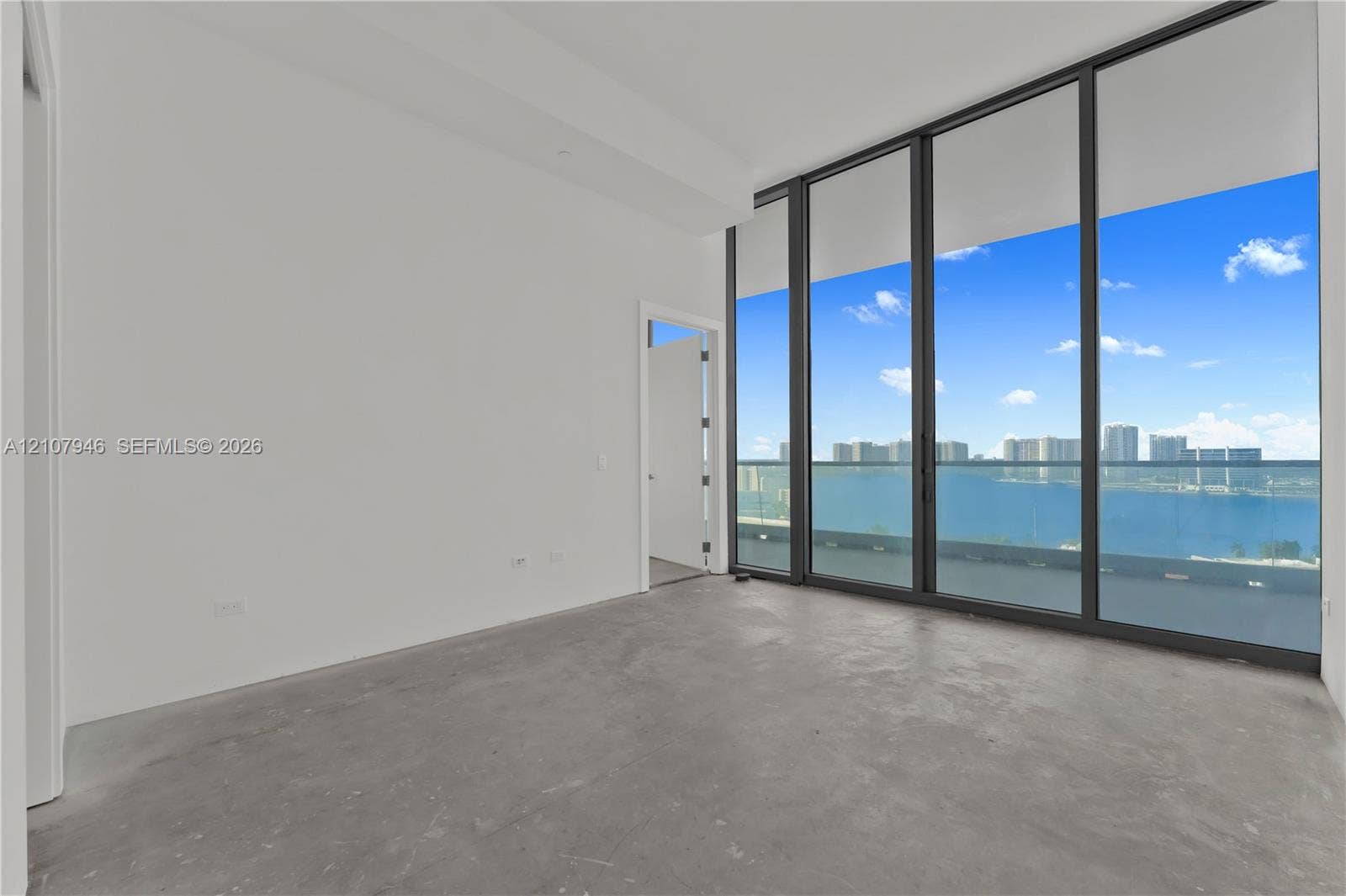 Floor-to-ceiling windows with black metal framing, concrete flooring, and access to an exterior glass-railed balcony with waterfront and skyline scenery