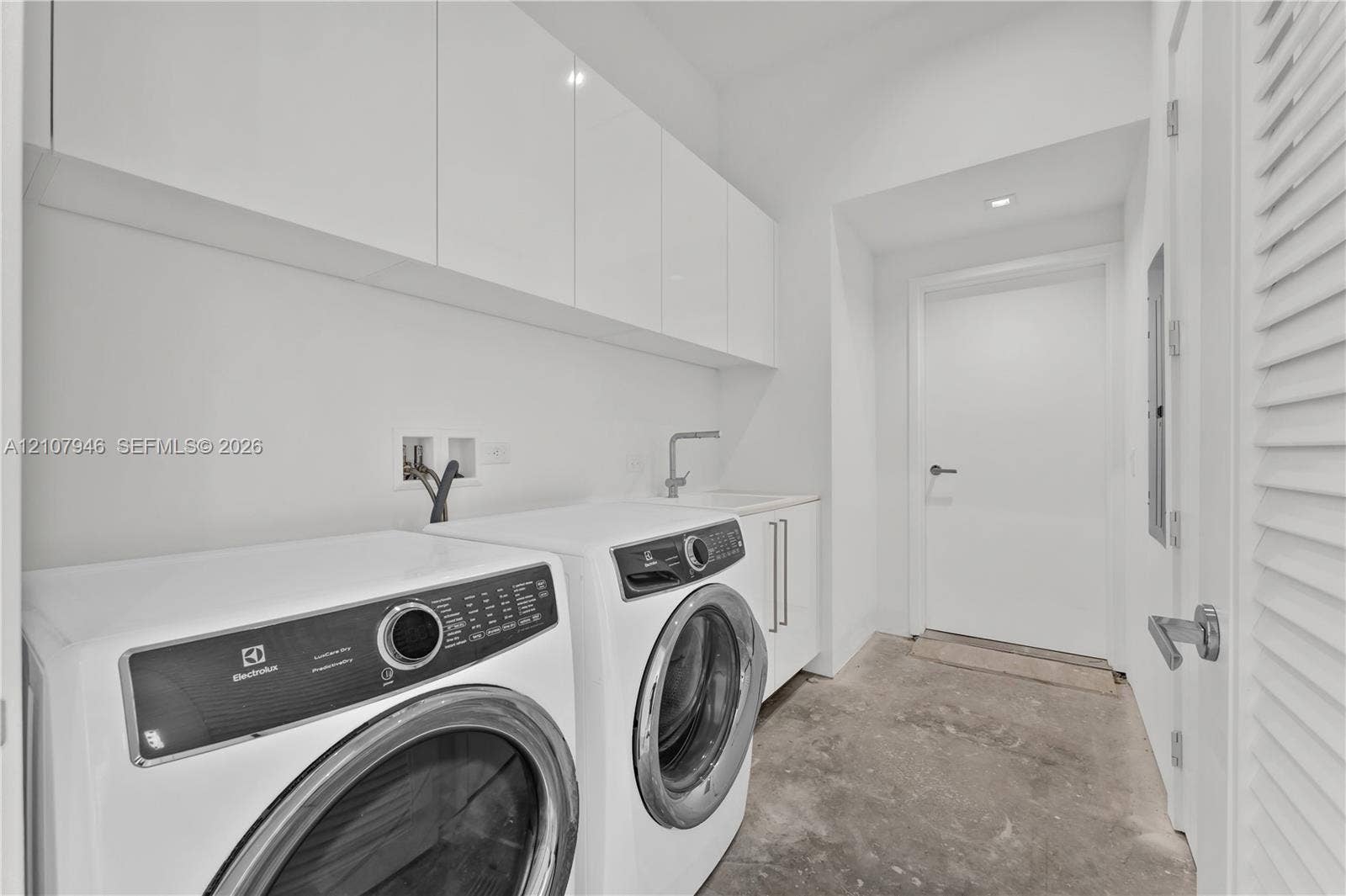 Laundry room featuring high-gloss white upper cabinetry, integrated sink with a chrome faucet, white base cabinetry, and concrete flooring