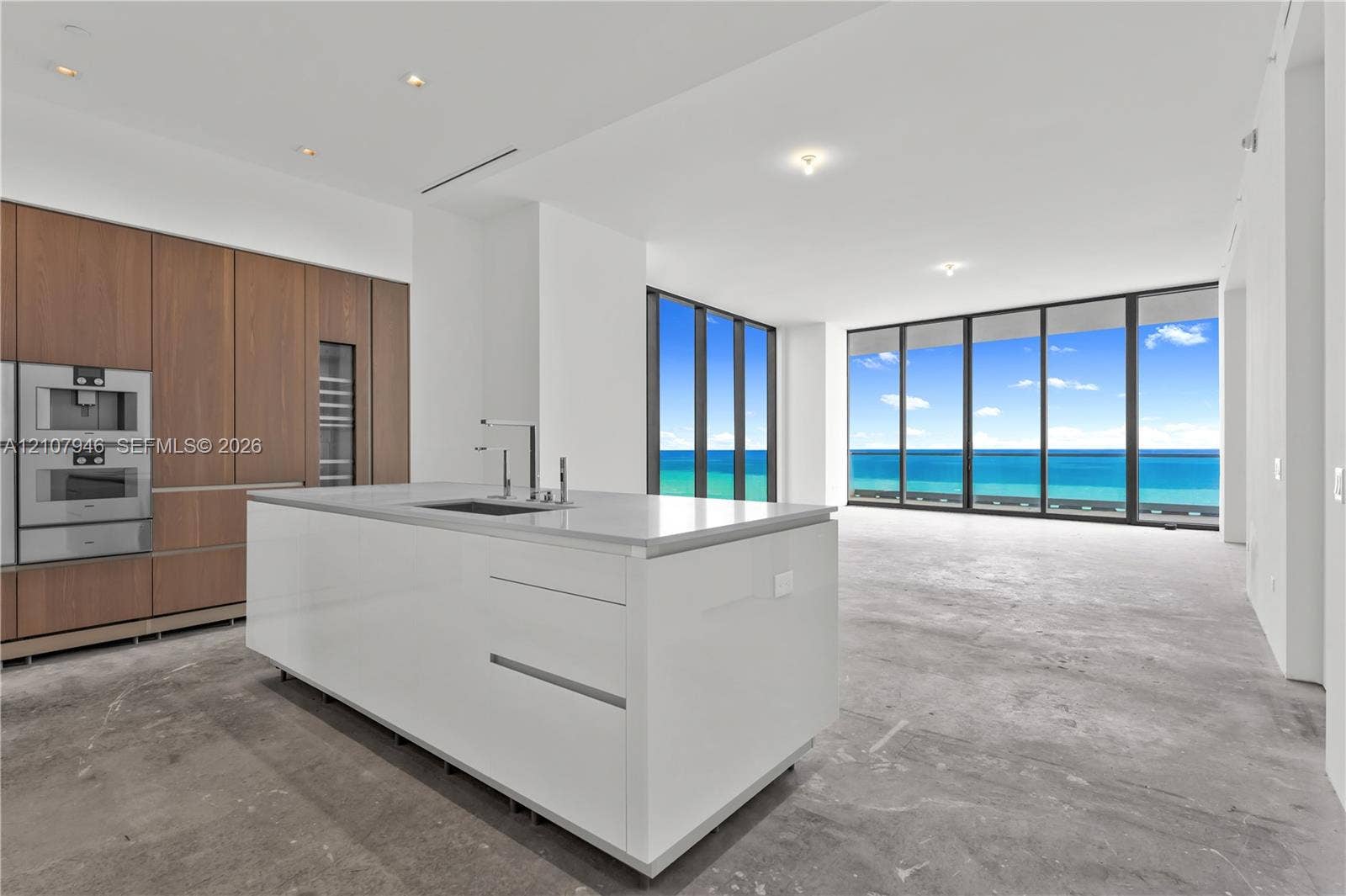 Kitchen featuring full-height wood-finish cabinetry, built-in stainless steel appliances, stone-surface center island with integrated sink, and floor-to-ceiling windows with ocean backdrop