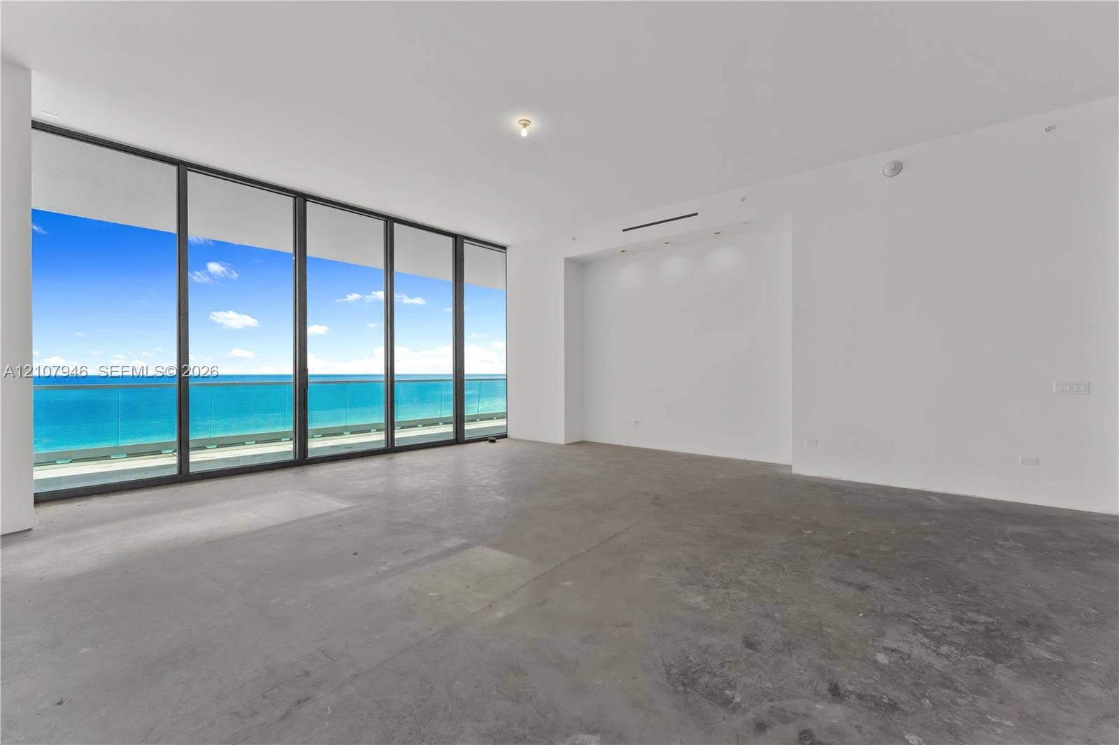 Floor-to-ceiling windows, full-height glass sliding doors, private glass-railed balcony, polished concrete flooring, and ocean backdrop