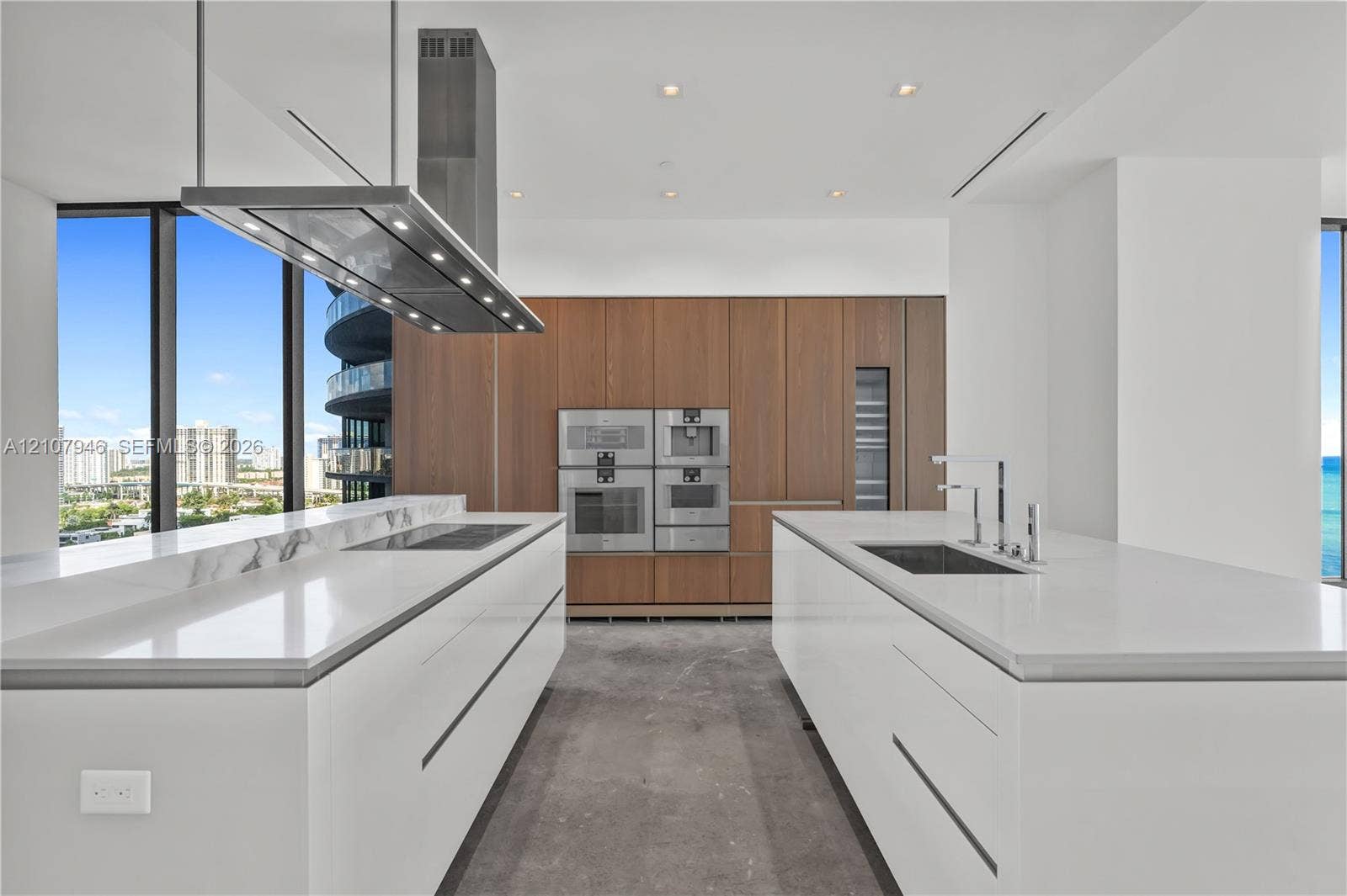 Kitchen featuring dual white islands with stone-topped surfaces, floor-to-ceiling wood-finish cabinetry, integrated stainless steel appliances, and a ceiling-mounted stainless steel vent hood
