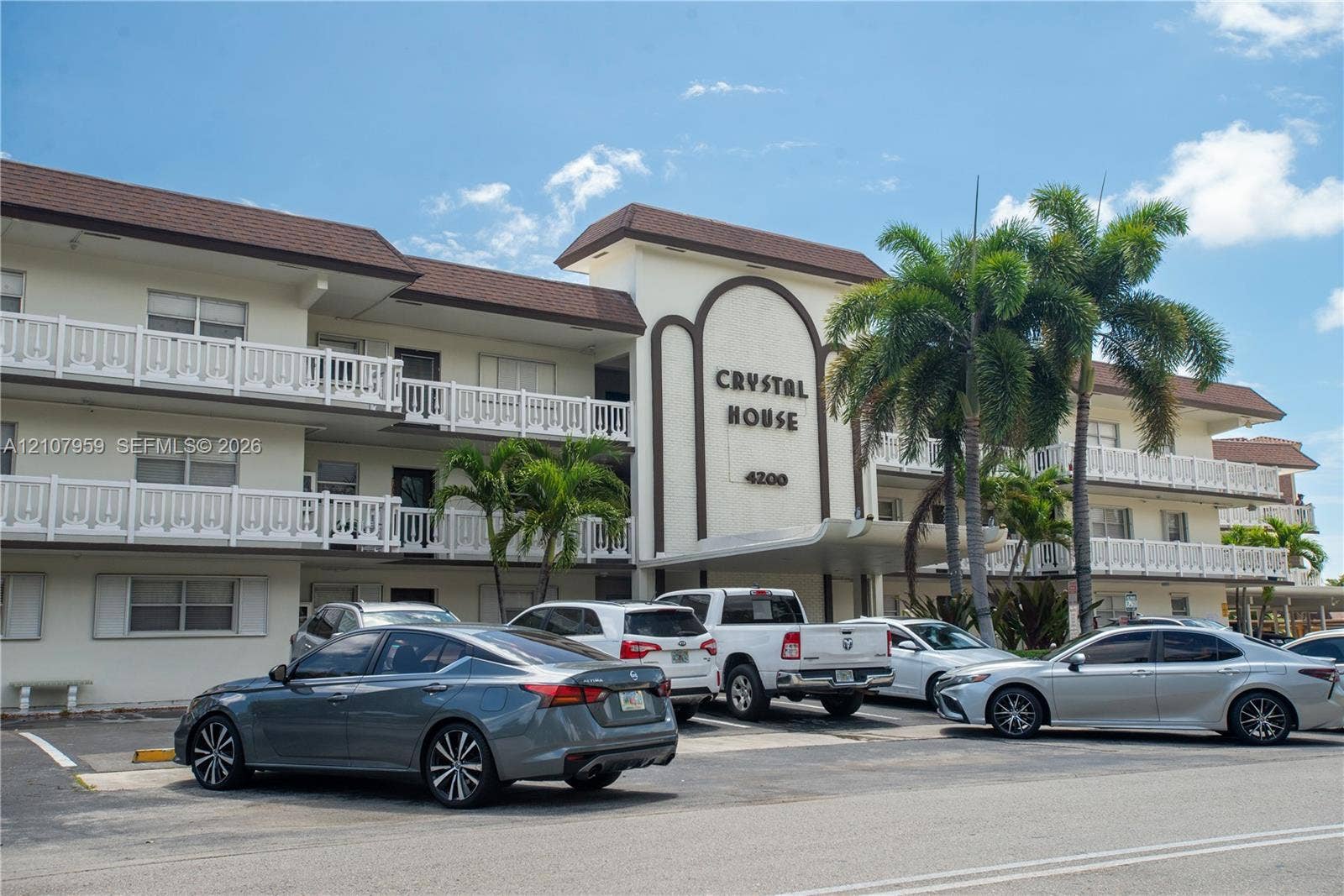 4200 Crystal Lake Drive, #204, Deerfield Beach, Deerfield Beach, FL 33064 Multi-story exterior featuring a shingled roof, white balcony railings, a covered entrance portico, and integrated mature palm landscaping