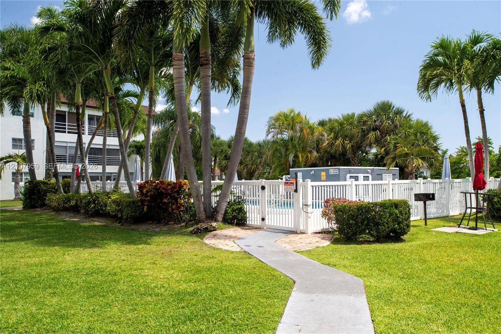 4200 Crystal Lake Drive, #204, Deerfield Beach, Deerfield Beach, FL 33064 Swimming pool area featuring a white vinyl perimeter fence, concrete paver pathway, manicured lawn, and mature palm trees