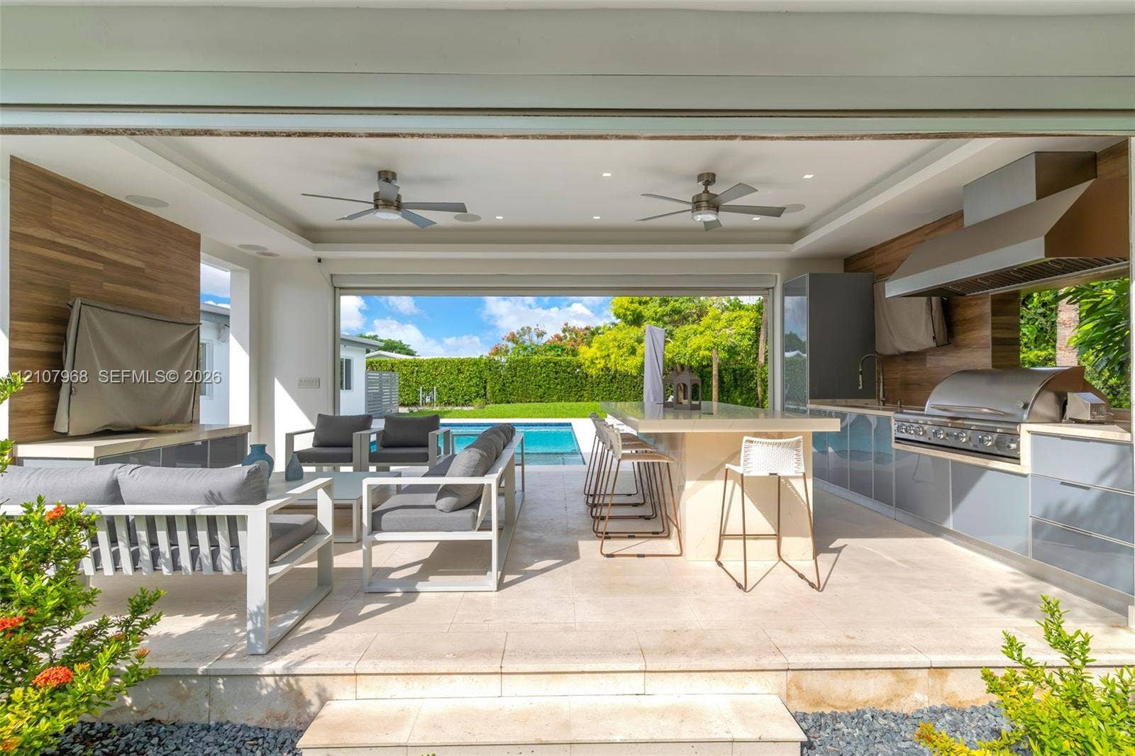 10841 SW 126th Street, Miami, FL 33176 Covered outdoor living space featuring a built-in kitchen with a professional-grade stainless steel grill, stone-topped island with seating, wood-finish wall paneling, and integrated ceiling fans