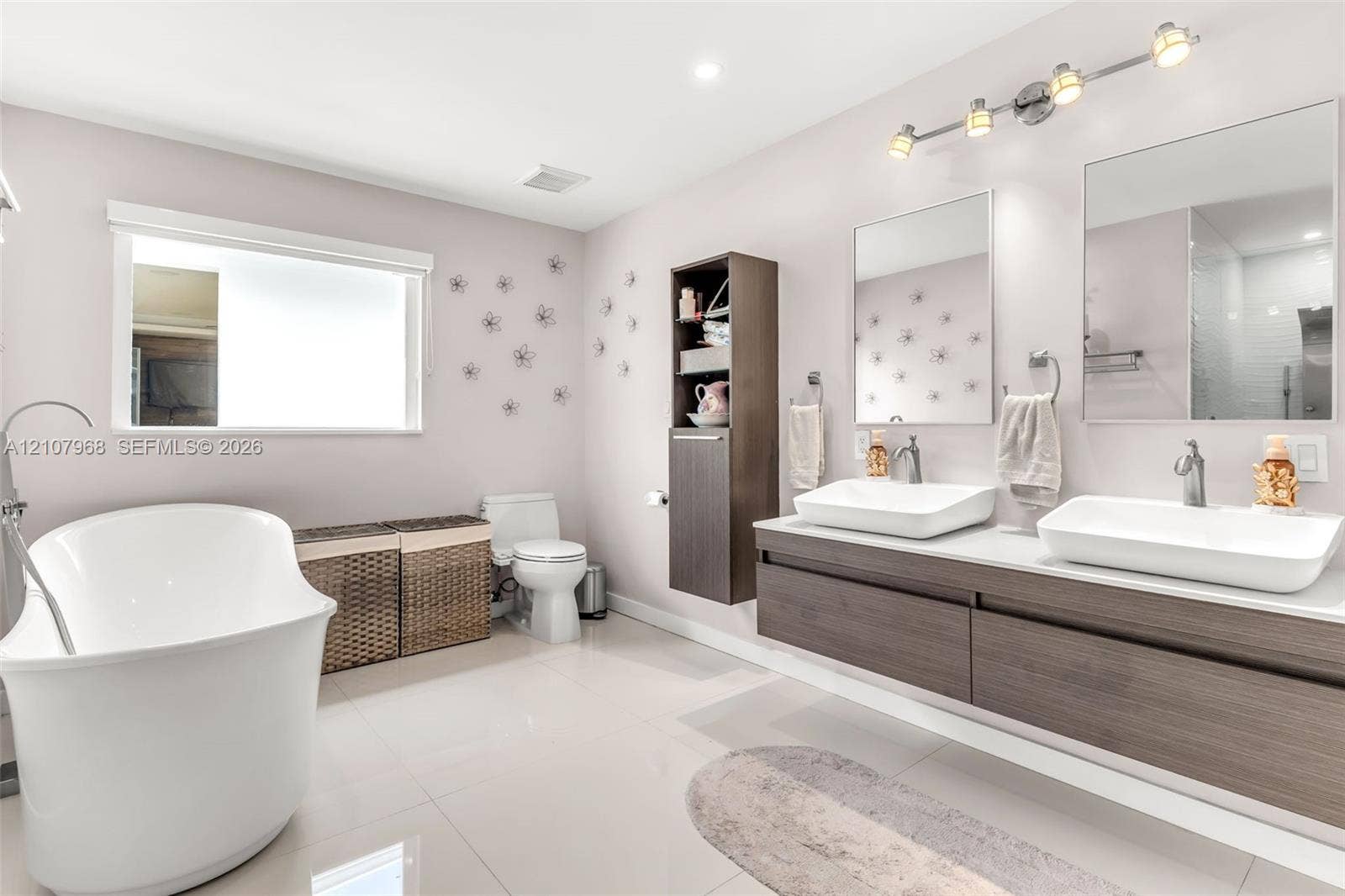 10841 SW 126th Street, Miami, FL 33176 Floating vanity featuring wood-finish cabinetry, dual vessel sinks, and stone-surface countertop, complemented by a freestanding soaking tub, vertical built-in storage cabinet, and rectangular wall-mounted mirrors