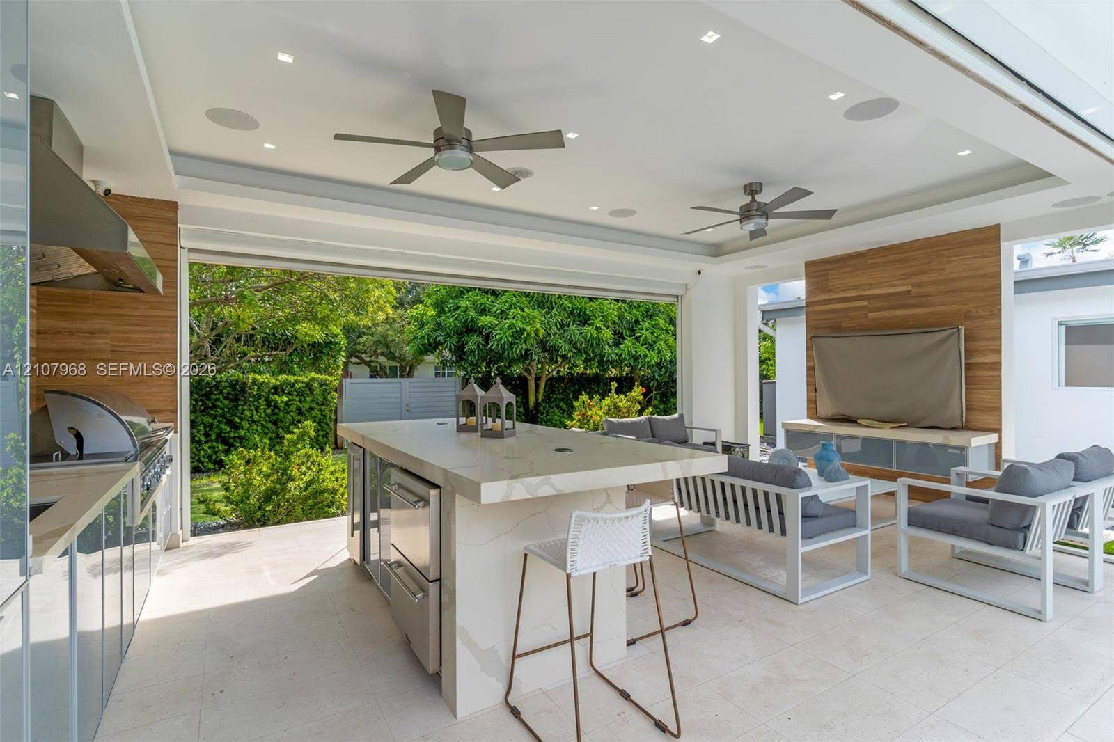 10841 SW 126th Street, Miami, FL 33176 Covered outdoor kitchen and living space featuring a stone-topped island with stainless steel appliances, wood-finish wall paneling, dual ceiling fans, and large-format tile flooring