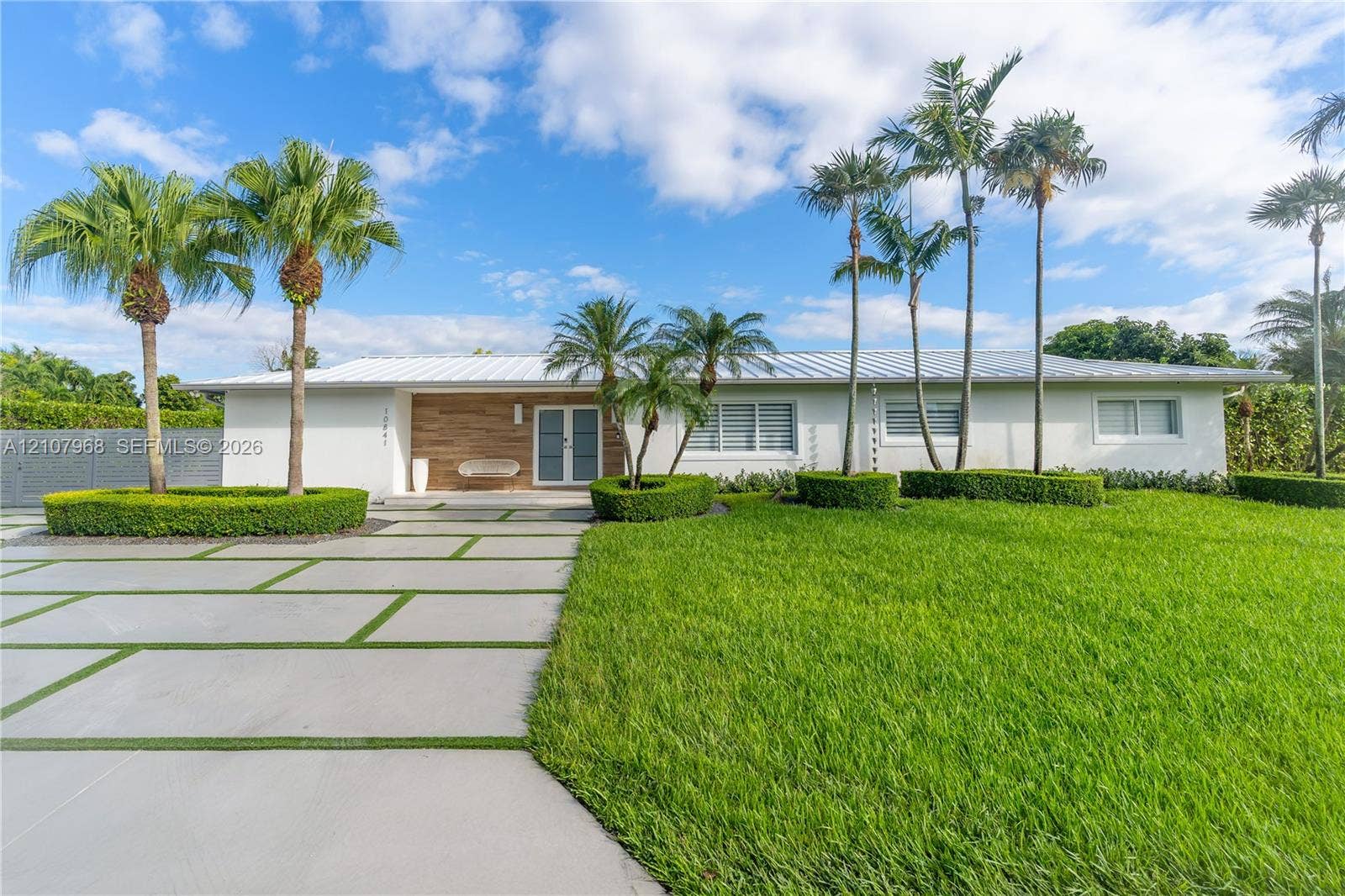 10841 SW 126th Street, Miami, FL 33176 Single-story residence featuring a standing-seam metal roof, horizontal wood-panel siding, a concrete paver driveway with integrated turf, and professional landscaping with mature palm trees