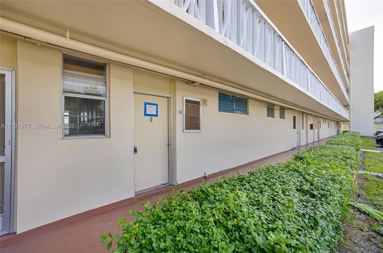 2910 Point East Drive, #M108, Aventura, FL 33160 Exterior walkway featuring a covered balcony, recessed window framing, solid-surface flooring, and mature hedge landscaping