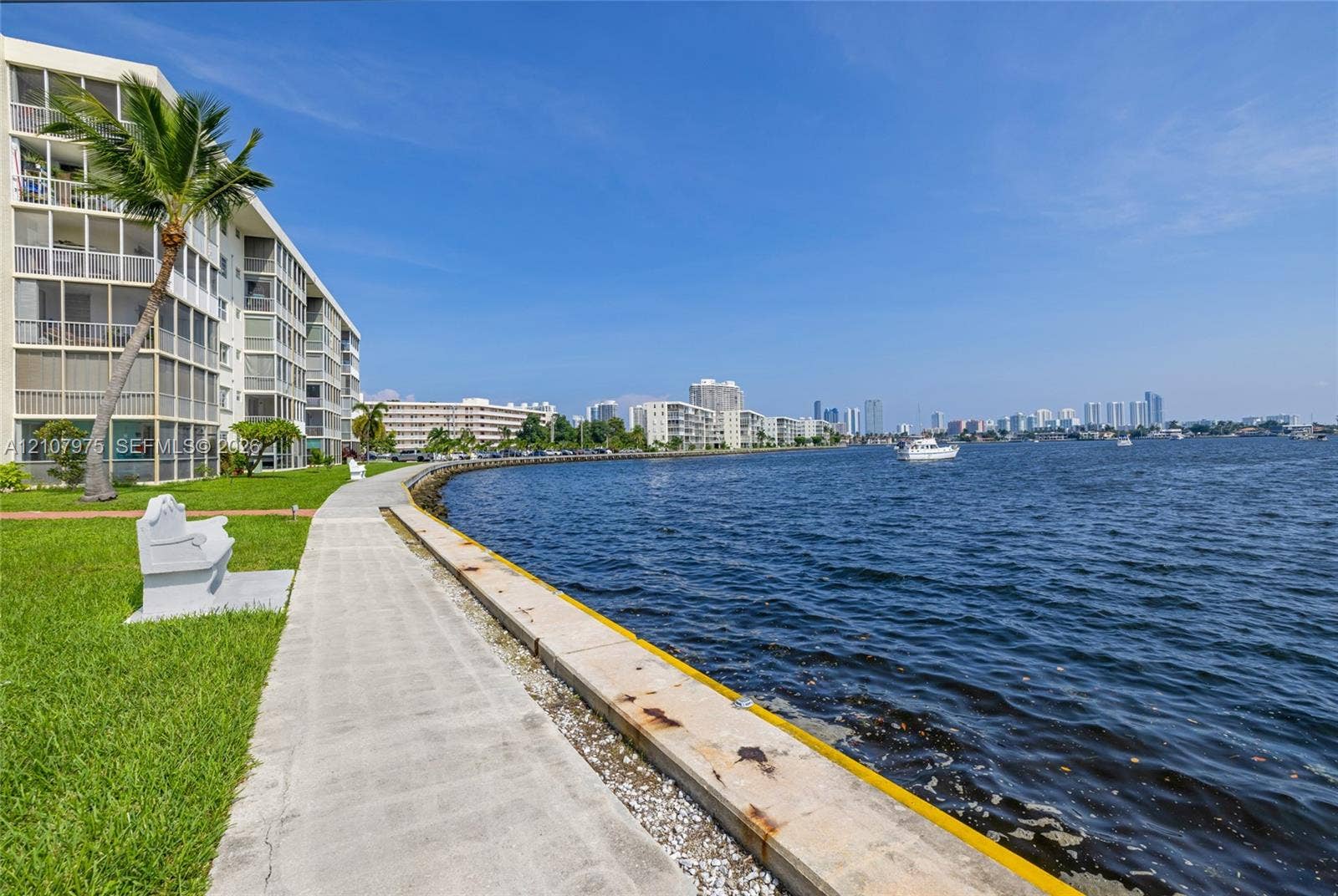2910 Point East Drive, #M108, Aventura, FL 33160 Waterfront multi-story residential building featuring screened-in balconies, concrete bulkhead, paved walkway, and manicured lawn