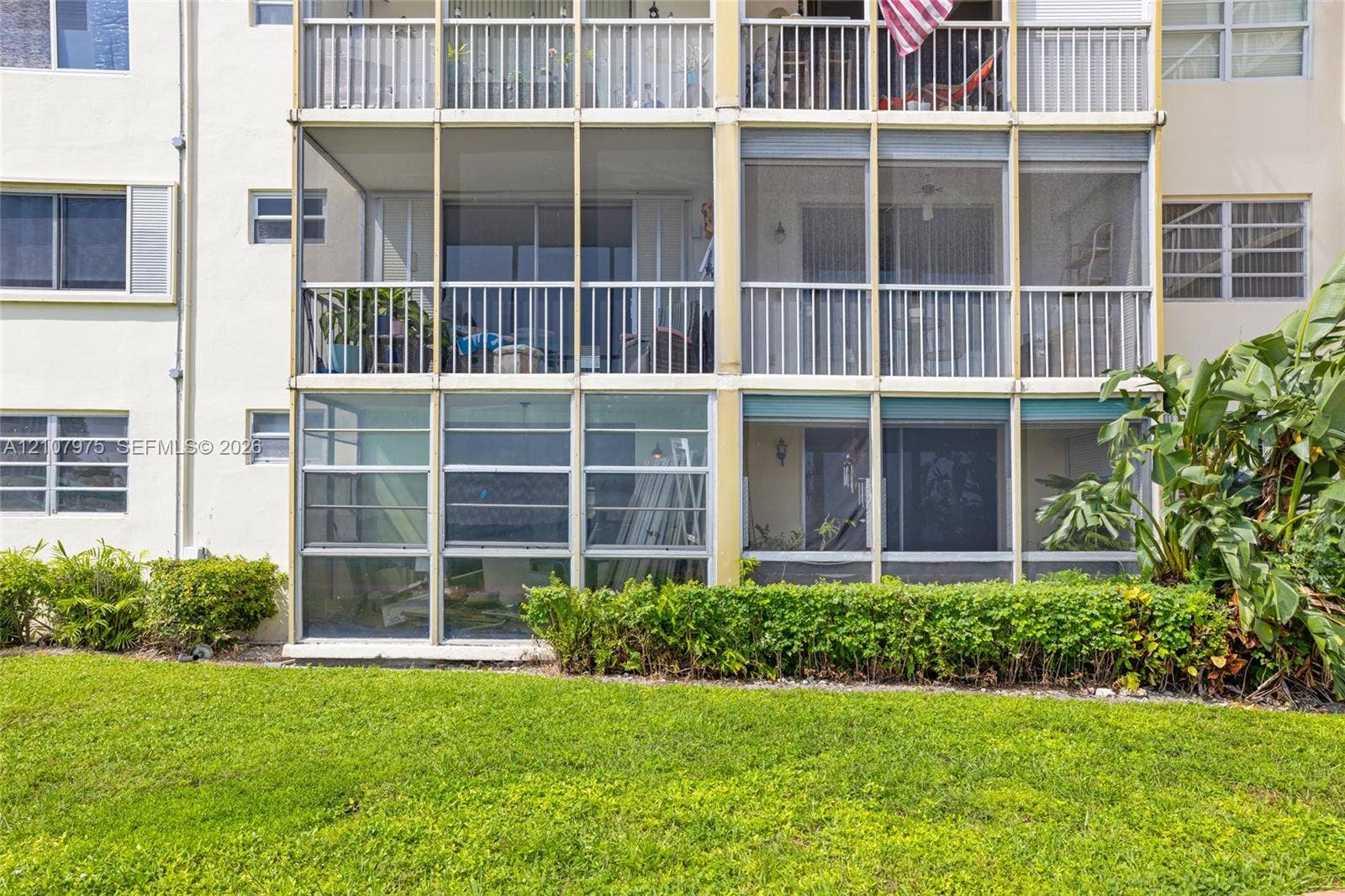 2910 Point East Drive, #M108, Aventura, FL 33160 Multi-story building exterior featuring screened-in lanais, aluminum-frame balcony railings, and exterior windows with horizontal sliding panes, situated on a manicured lawn with mature landscaping