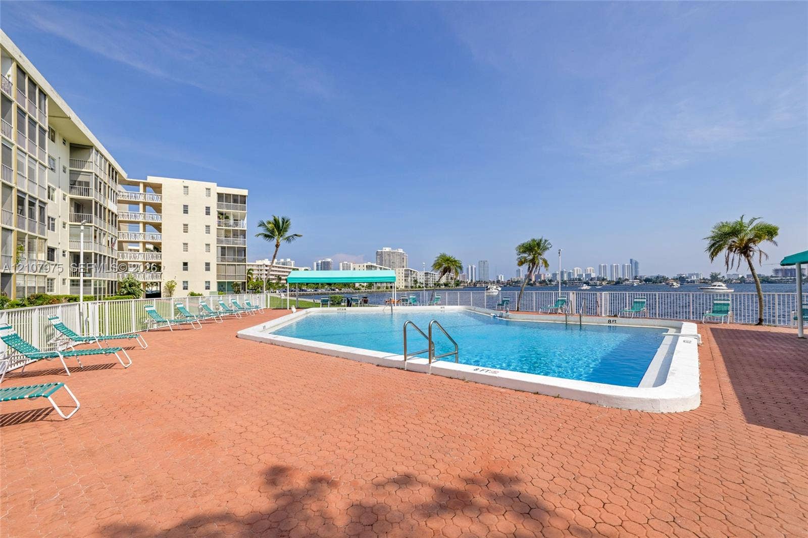 2910 Point East Drive, #M108, Aventura, FL 33160 Outdoor swimming pool with integrated metal ladder, surrounding brick-paver deck, protective perimeter fencing, and established palm trees