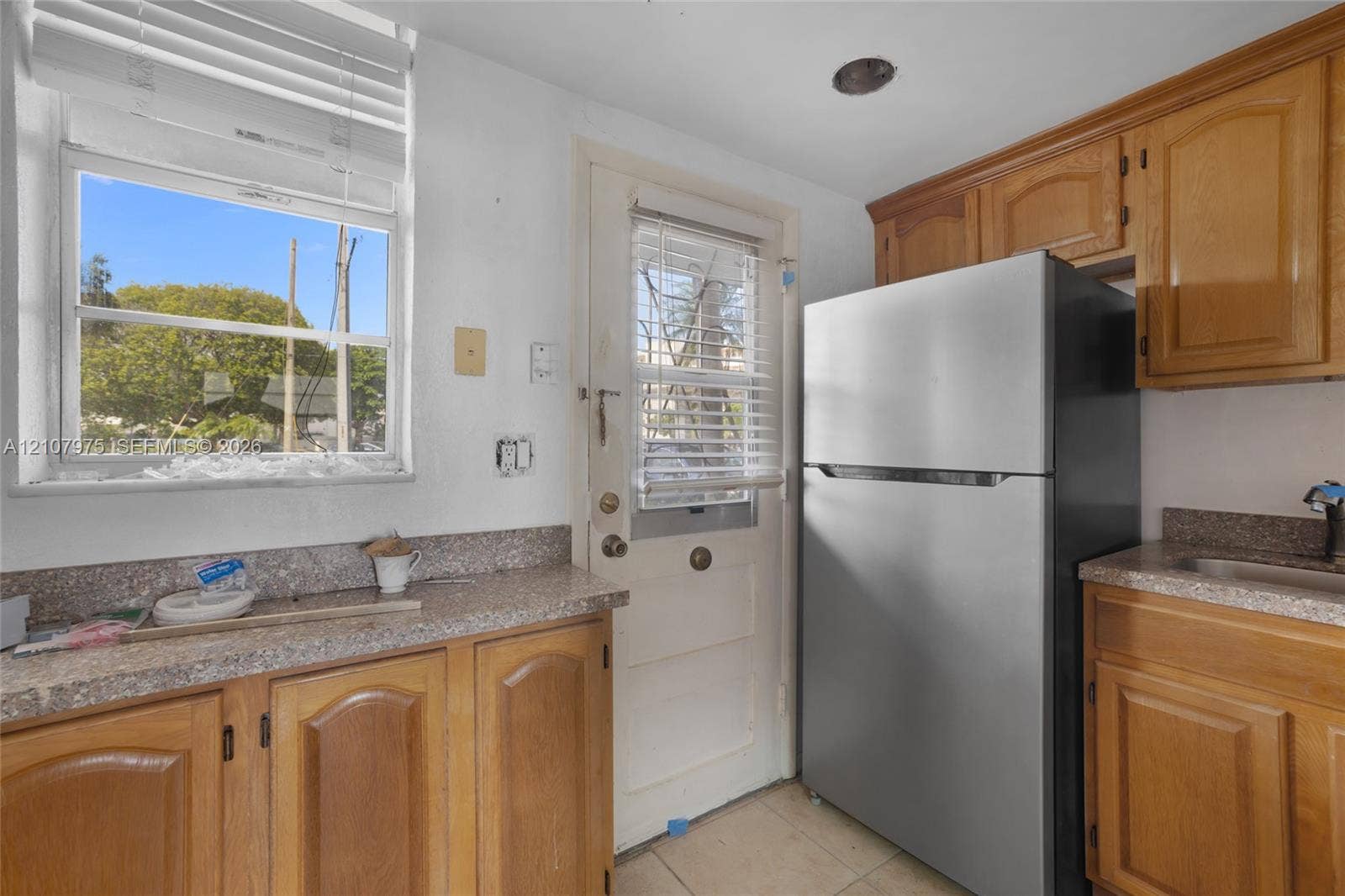 2910 Point East Drive, #M108, Aventura, FL 33160 Kitchen featuring solid wood cabinetry, stone-finish countertops, stainless steel refrigerator, and a dedicated entry door with integrated window blinds