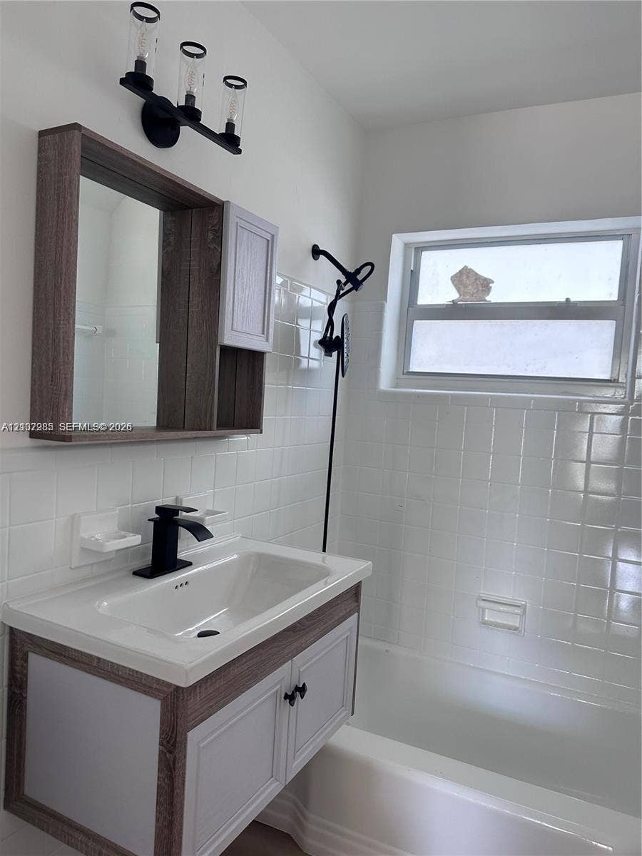Bathroom featuring a wood-finish vanity with a rectangular sink, matte black faucet, wall-mounted three-light fixture, and white tiled wall surround