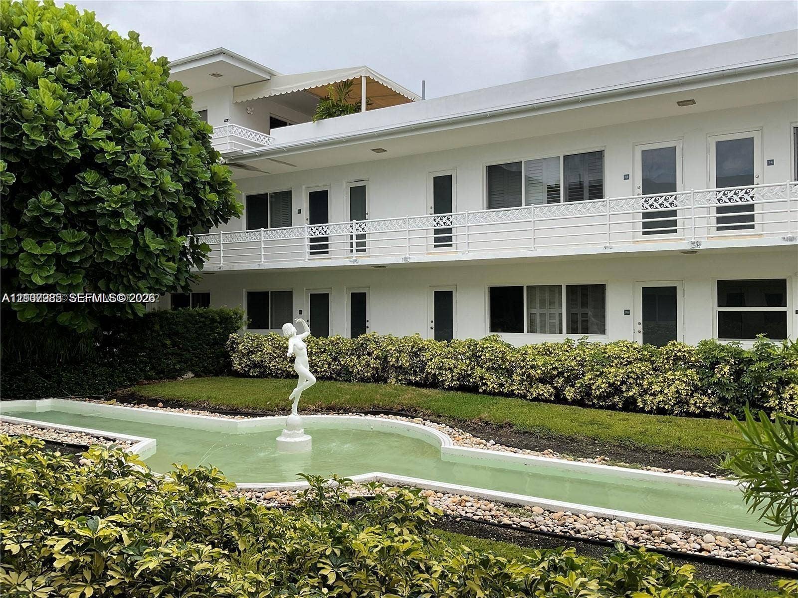 Exterior facade featuring white stucco walls, multi-level balconies with ornamental white railings, and a ground-level decorative water feature with a central sculpture surrounded by landscaped shrubbery