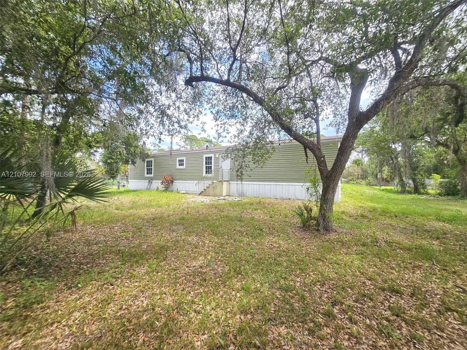 725 S Mayoral Street, Clewiston, FL 33440 Exterior featuring olive-toned horizontal siding, white trim, multiple windows, and a grassy lot with mature trees